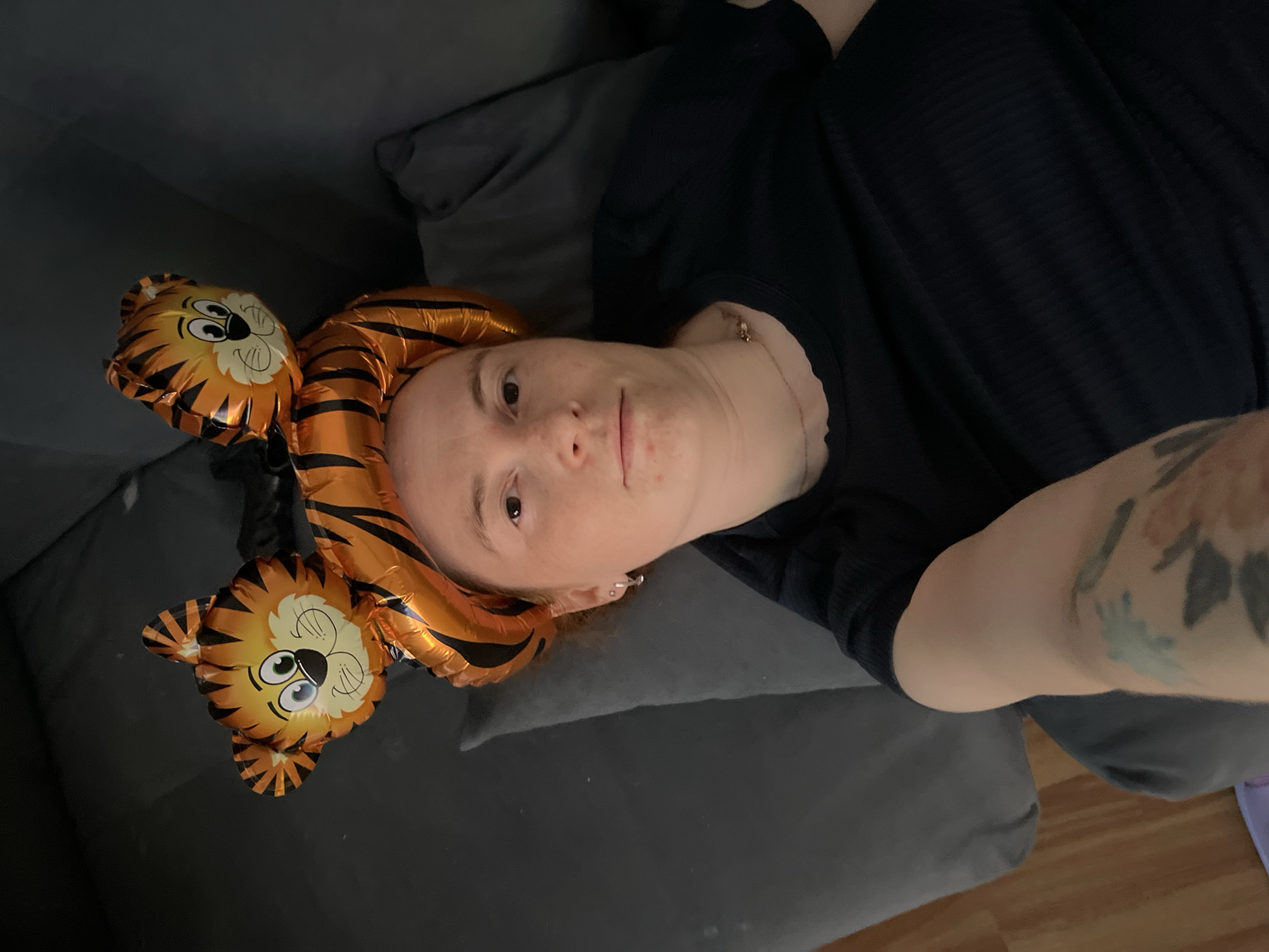 A selfie depicts a fair-skinned person lying on a gray couch or cushion. The person is wearing a black shirt. A tiger-themed balloon is positioned on the person's head. The person's arm, bearing a tattoo, extends into the frame. A glimpse of a wooden floor is visible. The location is unidentified beyond the general image context. There is no verifiable indication of Chaikovsky, Russia.