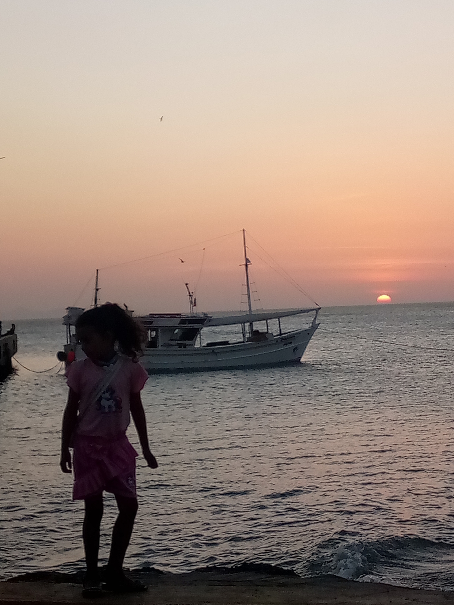 The image depicts a young girl in pink clothing standing on a concrete pier. Behind her is a boat anchored in the water. In the background, the sun is setting over the ocean. The image was taken at Juan Griego, Venezuela.