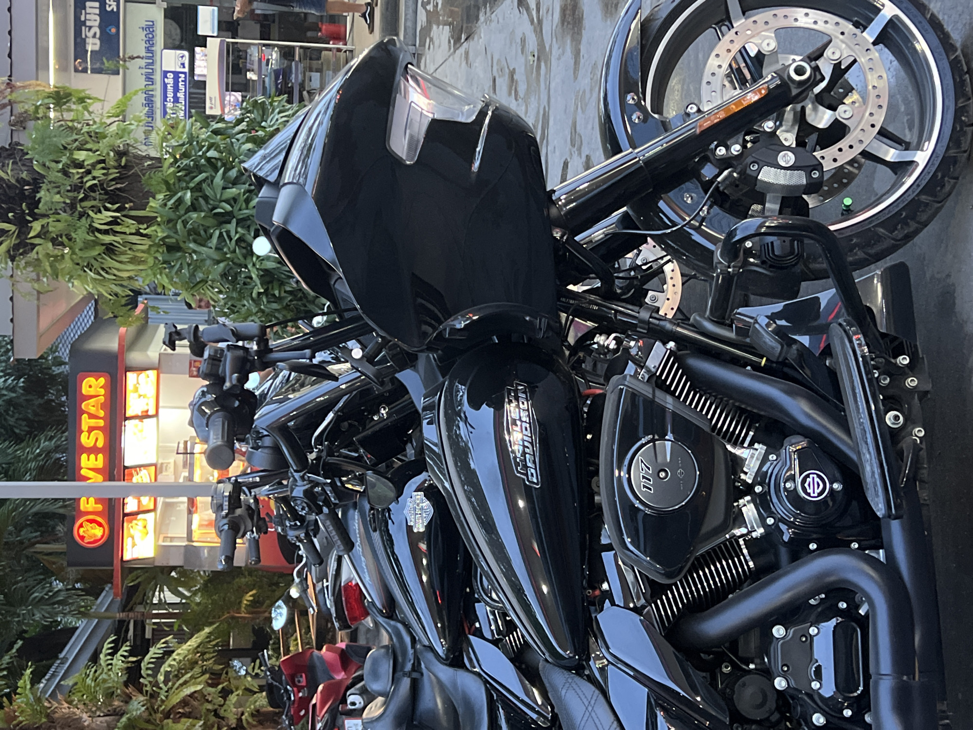 The image shows a row of black Harley-Davidson motorcycles parked in front of a "FIVE STAR" establishment, likely a food or retail business. The foreground features a detailed view of the bikes, highlighting the "HARLEY-DAVIDSON" logo, "117" engine details, and front wheel. In the background, there is a gas station sign in an East Asian language, along with greenery, suggesting the scene is located in a city in Southeast Asia.