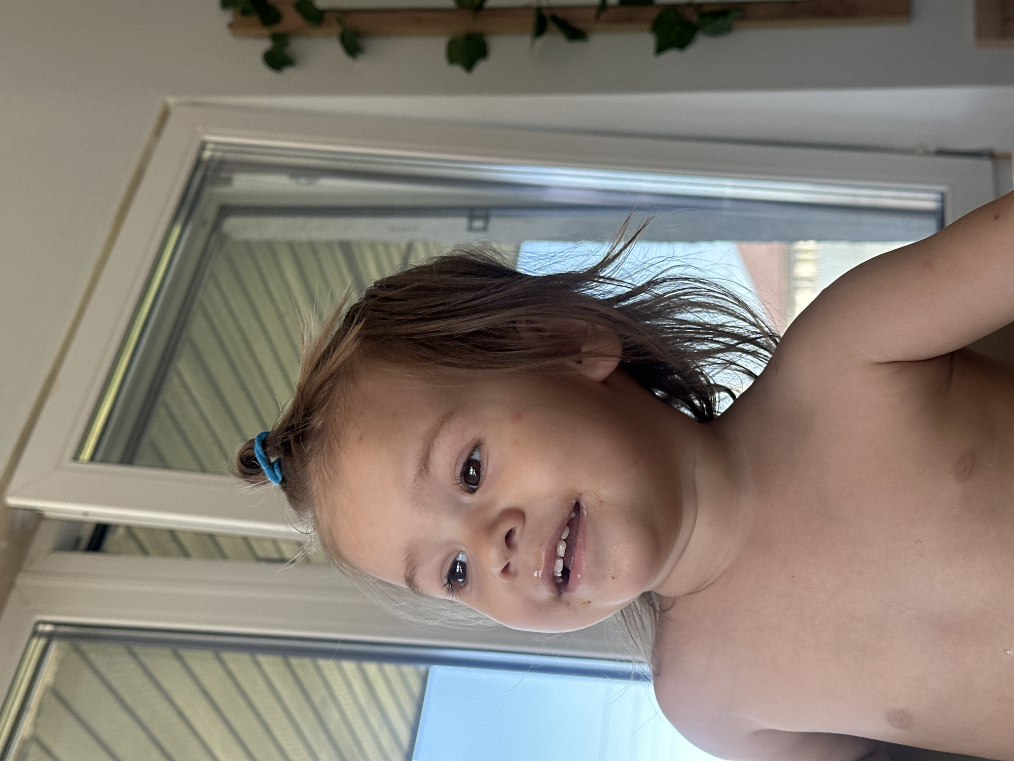 A young child with brown hair pulled back in a blue hair tie is smiling. They appear to have recently eaten, as evidenced by residue around their mouth. The child is shirtless. Behind the child is a window with a view of blinds or a slatted fence. Above the window, there is a wooden plank with artificial green vines. The location and city are not identifiable.
