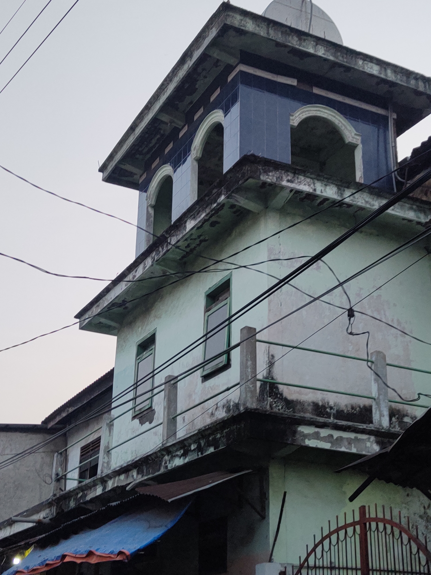 The image shows the corner of a light green, multi-story building with a weathered facade, possibly a mosque or religious structure, in Bangil, Indonesia. The building has several windows, some with green frames, and a prominent upper section decorated with blue tiles and arched openings. Overhead, a web of black electrical wires crisscrosses the frame. Below the building, there is a glimpse of a blue tarp and a dark metal fence. The sky is overcast and pale. The structure appears somewhat aged, with peeling paint and signs of wear.