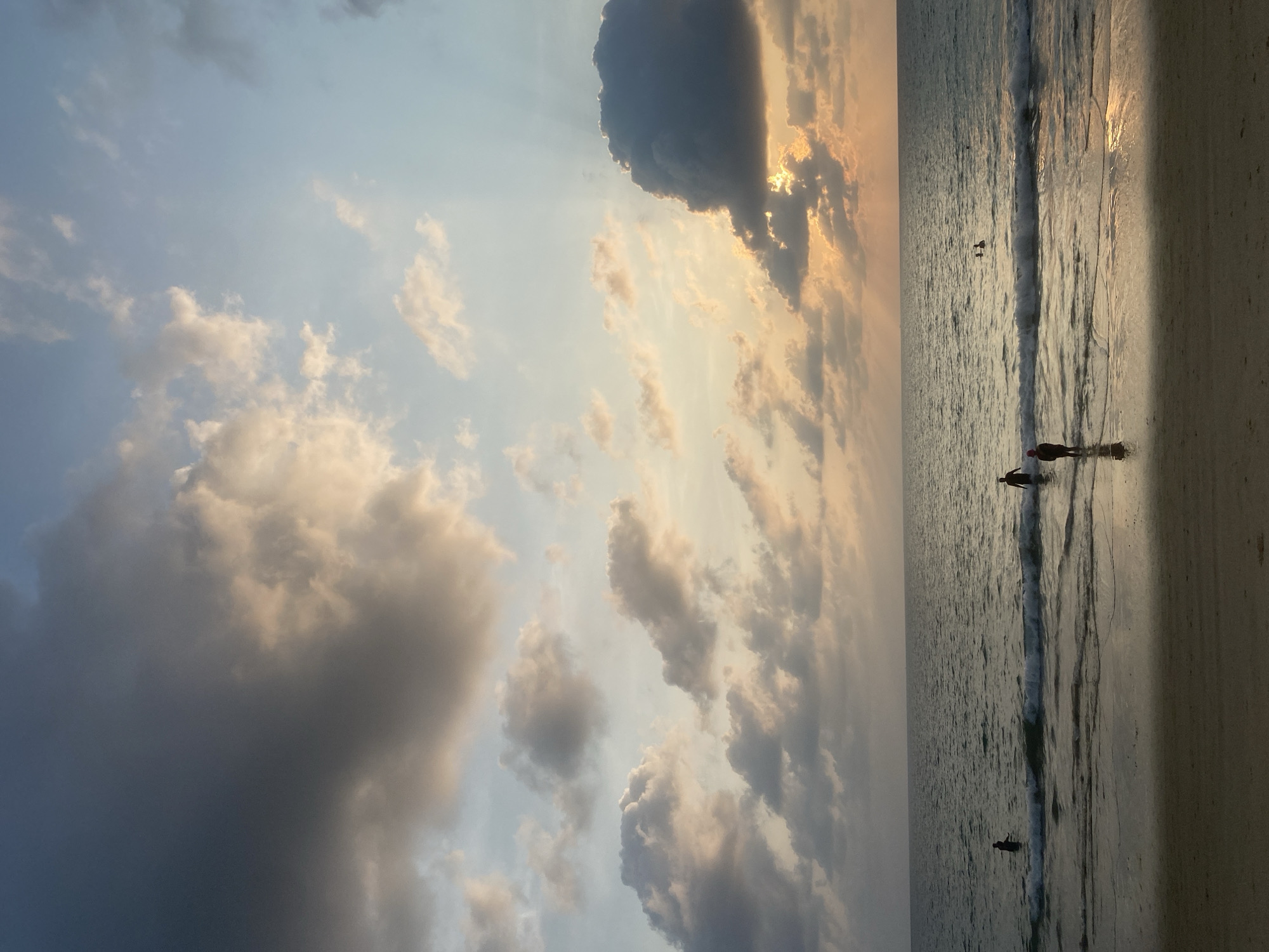 This media file captures a serene beach scene at either sunrise or sunset in Choeng Thale, Thailand. The sky dominates the upper portion, featuring a mix of dramatic, dark clouds and softer, lighter clouds illuminated by the low-hanging sun. Golden rays pierce through the clouds in the upper right, casting a warm glow across the horizon and reflecting on the calm ocean surface.
The setting is a tranquil beach, with the ocean meeting a wet, sandy shore. Several individuals are visible as dark silhouettes against the reflective water and sky. Two people are standing in the shallow water near the shore, appearing to wade or swim. Further out, another faint silhouette suggests someone is in the deeper water, possibly on a board or swimming. Gentle waves break along the shoreline, creating white foam.
The time of day is clearly indicated by the soft, golden light and the long shadows/silhouettes, suggesting either early morning or late evening. The weather appears calm, with a dynamic cloudscape. No text is visible in the image. The overall mood is peaceful and reflective, highlighting the natural beauty of the coastal environment at dusk or dawn.