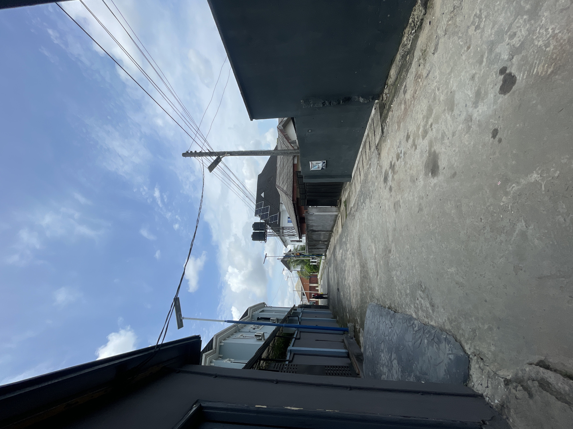 The image depicts a narrow concrete street or alleyway in Port Harcourt, Nigeria, captured during the daytime under a bright blue sky dotted with scattered white clouds. The perspective looks along the length of the street, with buildings flanking both sides.
In the immediate foreground and running down the right side of the street is a tall building with a dark grey, possibly black, facade. A small, white rectangular sign with unreadable text is affixed to its wall. Opposite, on the left side, another dark-colored building is partially visible, with its roofline at the bottom of the frame.
Overhead, multiple utility poles with a dense network of black wires crisscross the sky, a common sight in urban areas. One prominent pole with a crossarm stands further down the street on the right.
Further into the distance, other buildings become visible. A notable light-colored (possibly light blue or grey) building features several solar panels and large black water tanks mounted on its roof, indicating self-sufficient energy and water solutions often found in the region. A blue pipe or railing is visible along this building's exterior. In the far distance, near a reddish-brown structure, a blurry figure that appears to be a person can be discerned.
The concrete ground surface shows signs of wear, including various dark stains and patches. The weather is clear and sunny, suggesting a typical day. No specific activity is clearly depicted beyond the presence of the urban infrastructure.