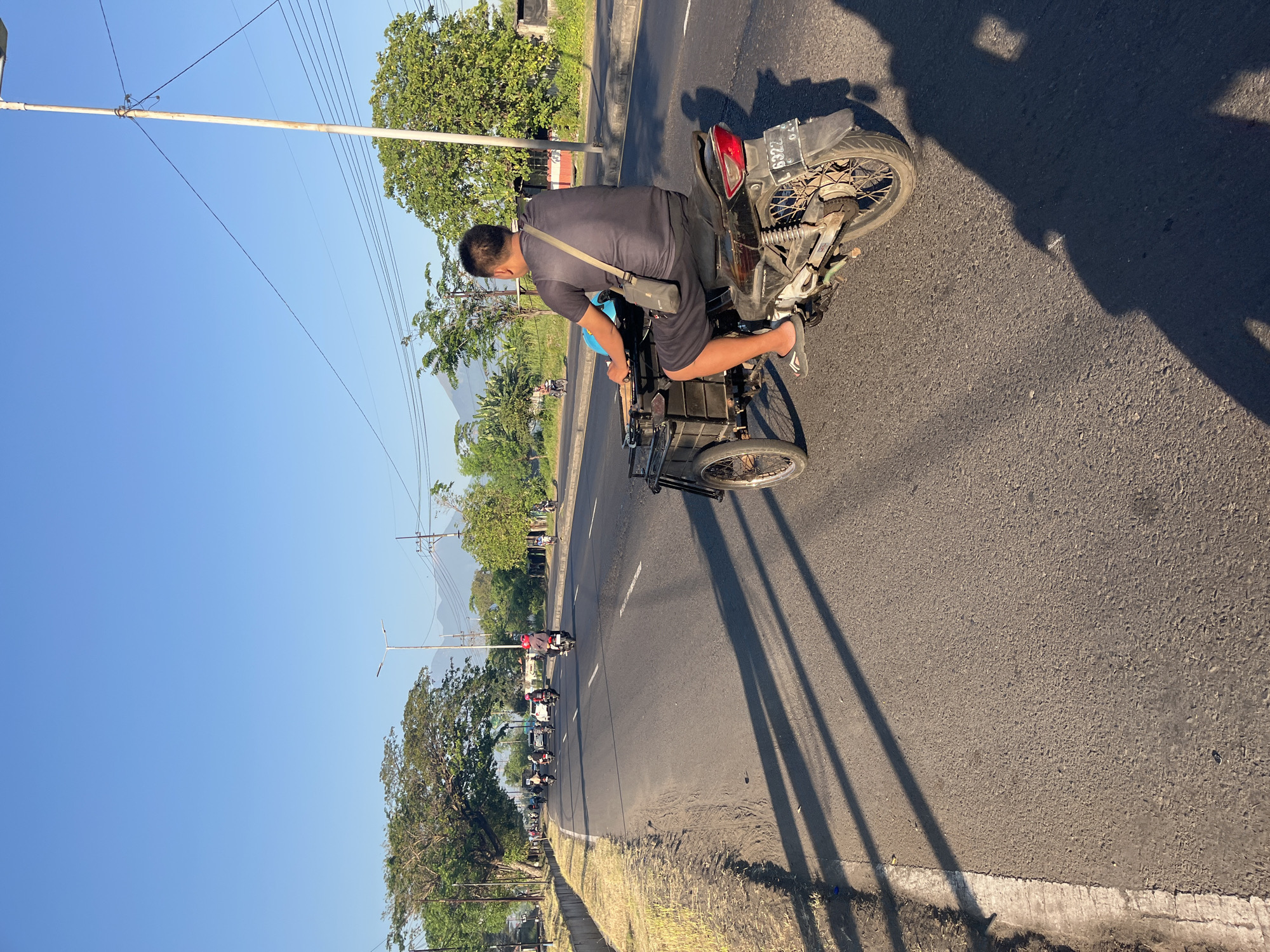 In Siring, Indonesia, a man is riding a motorcycle with a sidecar on a paved road. He is wearing a dark gray t-shirt and black shorts, with a bag slung across his chest. The motorcycle is black and has a license plate that reads "L 6322 04". The sidecar appears to be made of dark material and is attached to the left side of the motorcycle. In the background, there are other vehicles on the road, trees, streetlights, and mountains in the distance. The sky is clear and blue, and the sun is casting long shadows across the road.