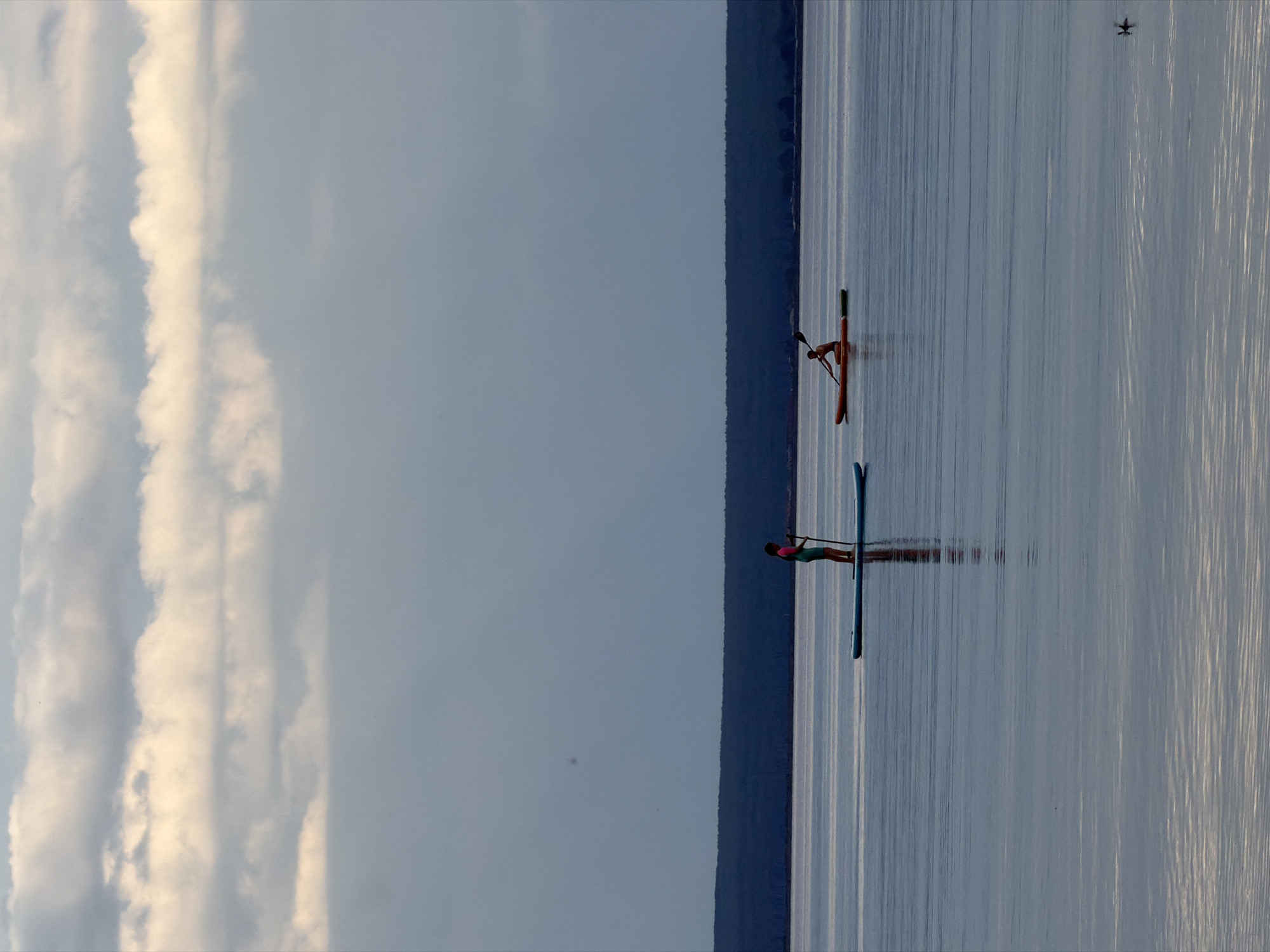Two individuals are engaged in water sports on a calm body of water. In the foreground on the left, a person is standing on a paddleboard and holding a paddle. To the right and slightly further back, another person is seated in a kayak and is paddling. The background features a dark, tree-lined horizon under a cloudy sky. This scene takes place on the Volga River near the village of Noviy, in the Ulyanovsk Oblast of Russia.