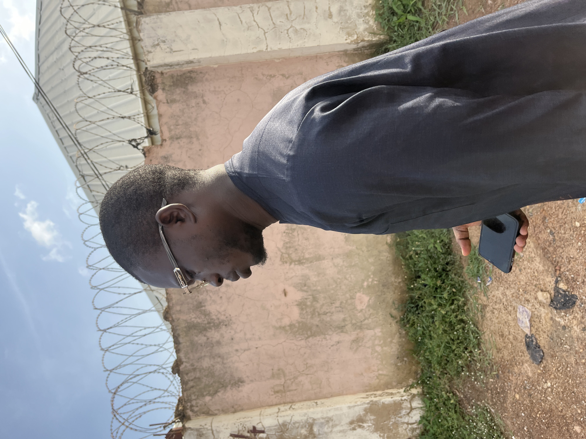 A dark-skinned man wearing glasses and a dark grey traditional outfit is seen in profile, looking downwards. He is holding a smartphone in his right hand. Behind him, a light pink wall with a rough texture is visible, topped with a coiled barbed wire fence. Beyond the wall, a corrugated metal roof of a building and the sky with scattered clouds can be seen. The ground in front of the wall is covered with dirt and patches of green grass. The scene appears to be an outdoor urban environment, possibly in Abuja, Nigeria, given the context.