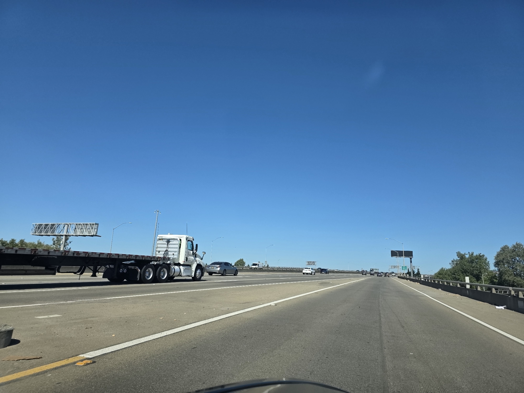 A wide-angle shot captures a bright, clear day on a multi-lane highway, likely in Sacramento, USA. The sky is a vibrant, cloudless blue, suggesting good weather. The road is asphalt, with distinct white lane markings and a yellow line on the left shoulder.
In the foreground, a flatbed truck with a white cab and a long, empty trailer is traveling in the right lane. The truck has multiple axles and large rear wheels. Further down the highway, several other vehicles are visible, including a silver sedan, a white car, and some cars in the distance. The scene appears to be during the daytime, with the sun casting shadows and illuminating the road.
To the left of the highway, a large billboard structure is visible, supported by a tall metal frame. On the right side of the road, trees line the median and the edge of the highway. The overall atmosphere is one of travel and transportation on a typical sunny day. There is no visible text in the image.