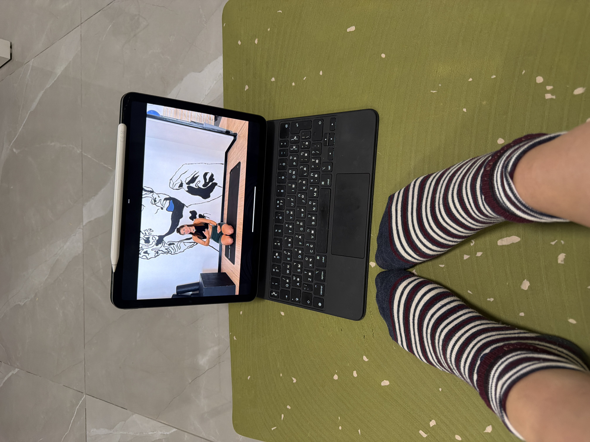 The image shows a first-person perspective of someone's legs and feet, positioned on a green exercise mat with light speckles. The person is wearing striped socks with navy, maroon, and white bands. To the left, a grey tiled floor is visible.
In the center, an open tablet (likely an iPad Pro) with a black Magic Keyboard case is resting on the mat. An Apple Pencil is magnetically attached to the side of the tablet. The tablet screen displays an image or video of a woman in athletic wear (black top, dark green shorts) performing what appears to be a fitness or yoga pose. She is in a room with a distinctive black-and-white mural on the wall featuring stylized hands and other abstract shapes. A dark, possibly wooden, fitness block or bench is beneath her.
The keyboard of the tablet is black and features a QWERTY layout with both English and Hebrew characters printed on the keys, indicating a bilingual keyboard. Standard function keys like "control," "option," "cmd," and "opt" are also visible. The scene suggests an indoor setting, potentially in Bat Yam, Israel, given the Hebrew keyboard and location context, where the individual is engaged in or preparing for a workout or relaxation session, using the tablet for content related to their activity.