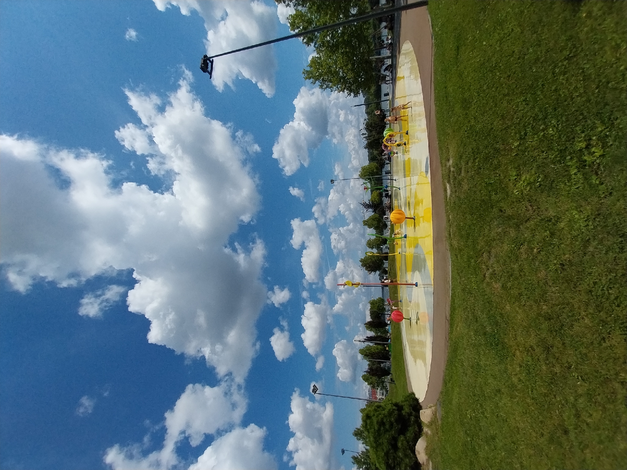 The image shows a splash pad in a park area, likely located in the city of Dnipro, Ukraine. The splash pad itself is a circular, flat surface with yellow and white sections and various water features. Several children are present, playing and interacting with the water spouts. In the background, there are trees, a body of water, and buildings. The sky is a vibrant blue with fluffy white clouds. A lamppost is visible on the right side of the image, and some grass and rocks are in the foreground.