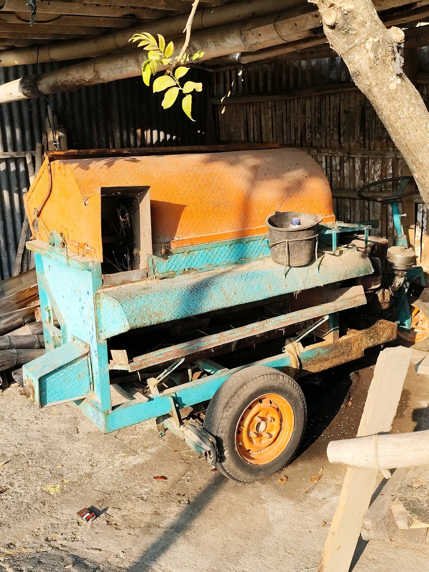 This image shows a brightly colored, aged agricultural machine, likely a thresher or similar processing unit, parked outdoors in a rustic, covered area. The machine has a large, orange, diamond-patterned hopper on top and a blue, diamond-patterned body with visible signs of rust and wear. It is supported by a teal-colored frame and has one visible orange wheel with a black tire, indicating it's a mobile unit. A black bucket sits on top of the machine. The background consists of corrugated metal siding and a wooden slatted wall, suggesting a utilitarian shed or workshop. A tree trunk leans into the frame from the upper right, and branches with green leaves hang from the ceiling structure. The ground is concrete, littered with debris, and bathed in strong sunlight casting shadows. The scene appears to be in Jebugan, Indonesia, given the context. There are no people present in the image.