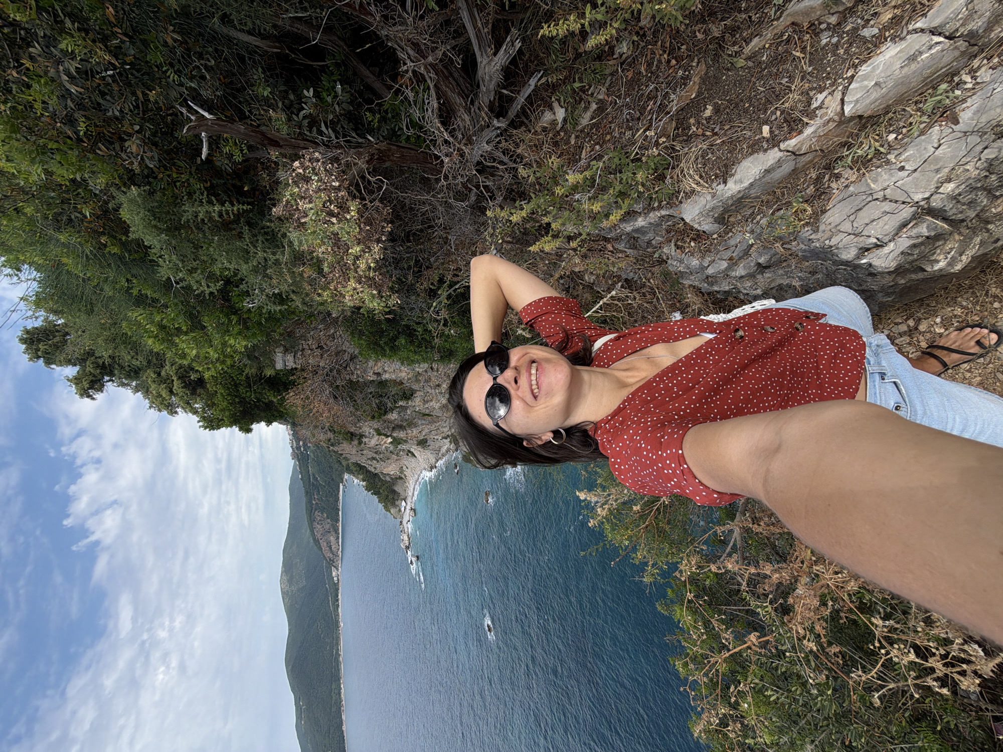 A cheerful woman is captured in a tilted selfie, seemingly reclining or lying down on a rocky cliffside overlooking the sea. She has dark hair and is wearing sunglasses pushed up onto her head, a red top with white polka dots, and light blue jeans. Her left arm is bent behind her head, and she is smiling broadly, revealing teeth.
The setting is a vibrant coastal landscape identified as Budva, Montenegro. In the background, a vast expanse of deep blue sea stretches out, dotted with a few small boats. A rugged coastline with green, treed cliffs extends into the distance, meeting a bright blue sky with scattered white clouds. The immediate foreground around the woman shows dry grass, rocks, and green shrubbery. The overall impression is one of leisure and enjoyment on a sunny day in a picturesque natural environment. There is no visible text in the image.