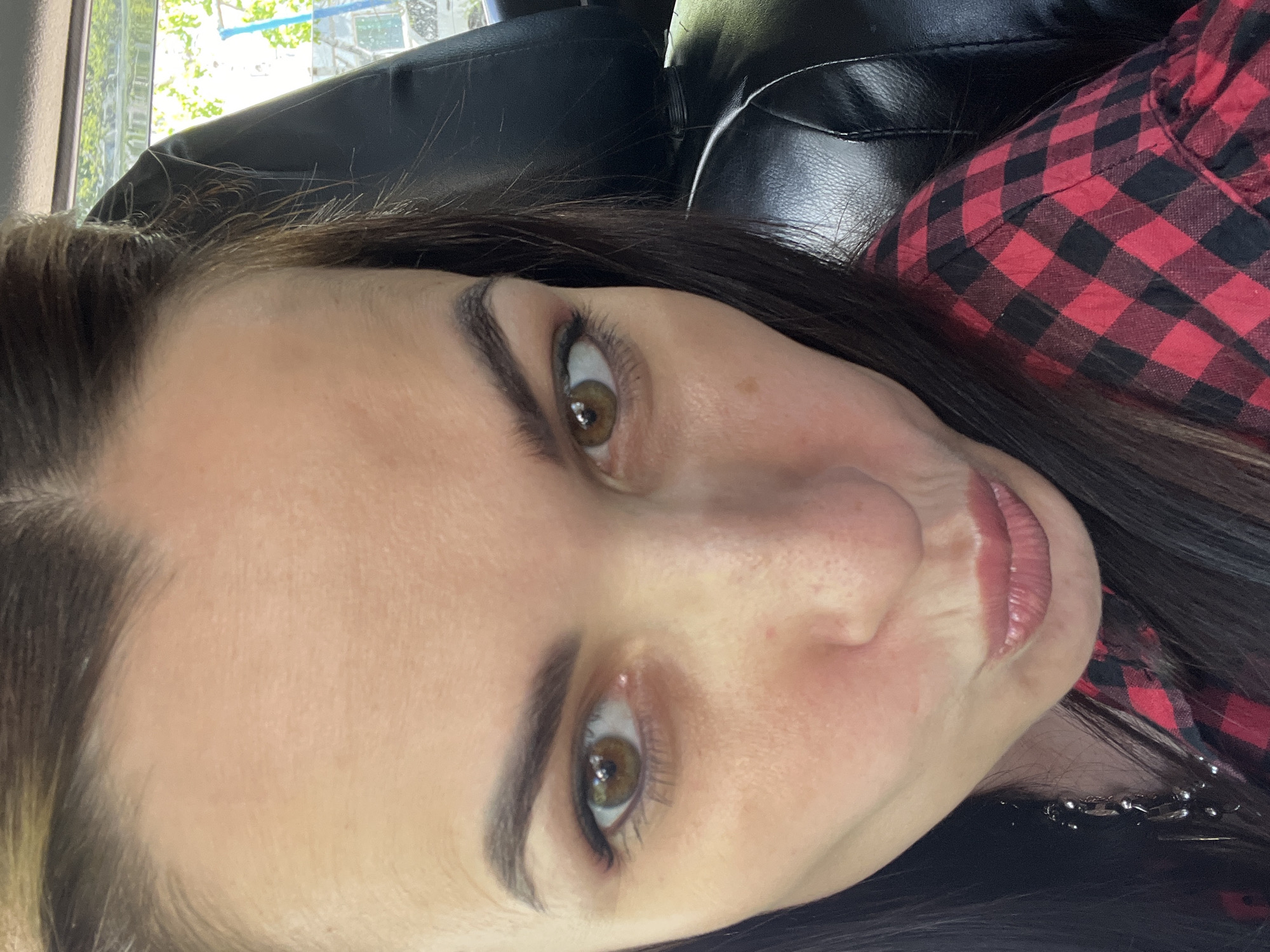 A close-up shot depicts a woman with dark hair and brown eyes, wearing a red and black plaid shirt. She is positioned in what appears to be a vehicle, indicated by the black leather interior visible in the background. The location is indeterminate based on the image.