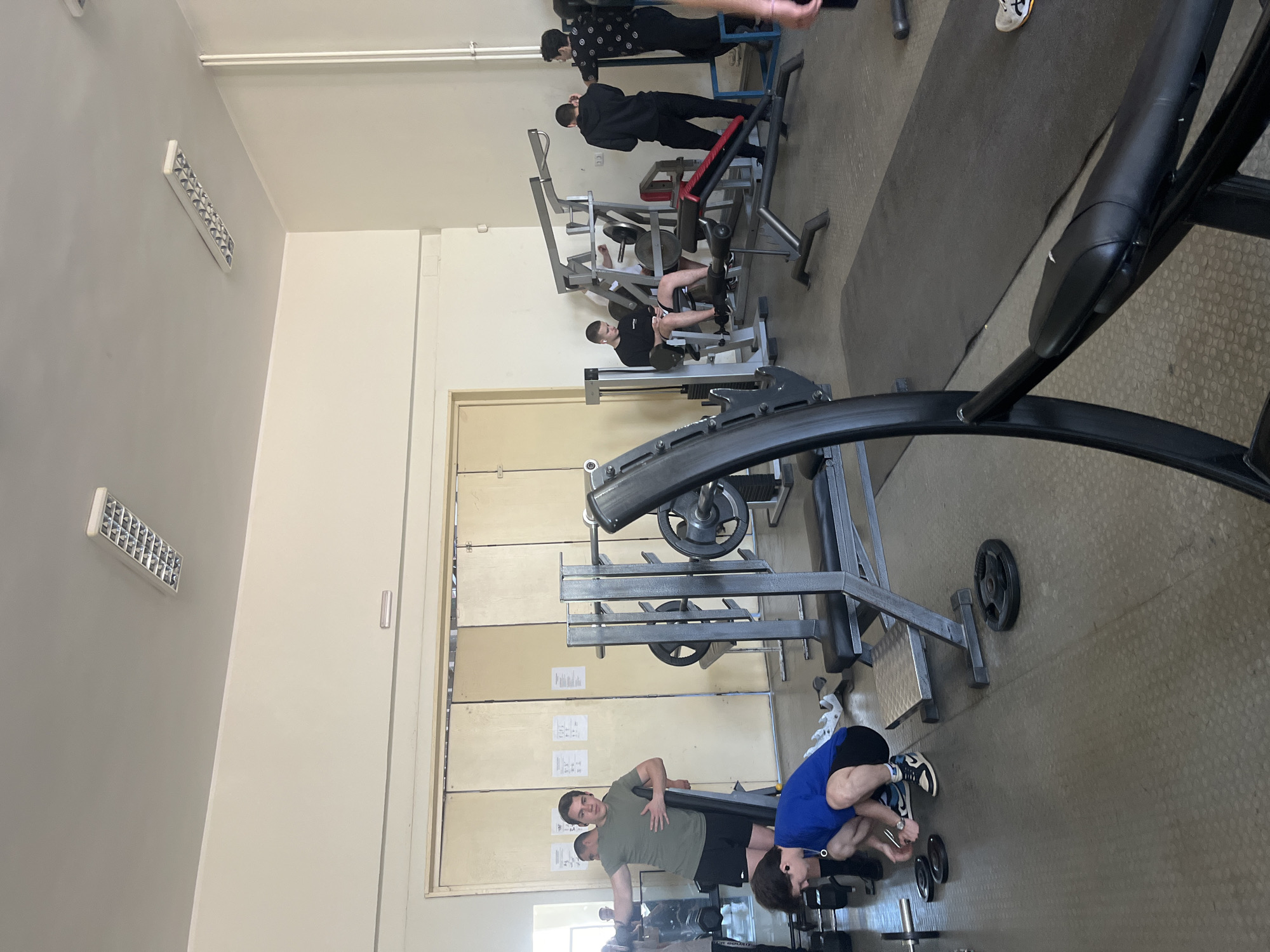 The image shows the interior of a gym. Several people are engaged in weightlifting activities using various weight machines. One person is bending over near a bench with dumbbells. Other individuals are using machines positioned throughout the gym space. The gym has typical equipment such as weight benches, leg press machines, and a rack with weights. The walls are painted a light color with rectangular fluorescent lights. There are large doors on one wall with informational posters. The floor is a neutral-colored surface. The scene is likely taking place in a fitness center. The location (Sofia, Bulgaria) is not discernible from the image content.