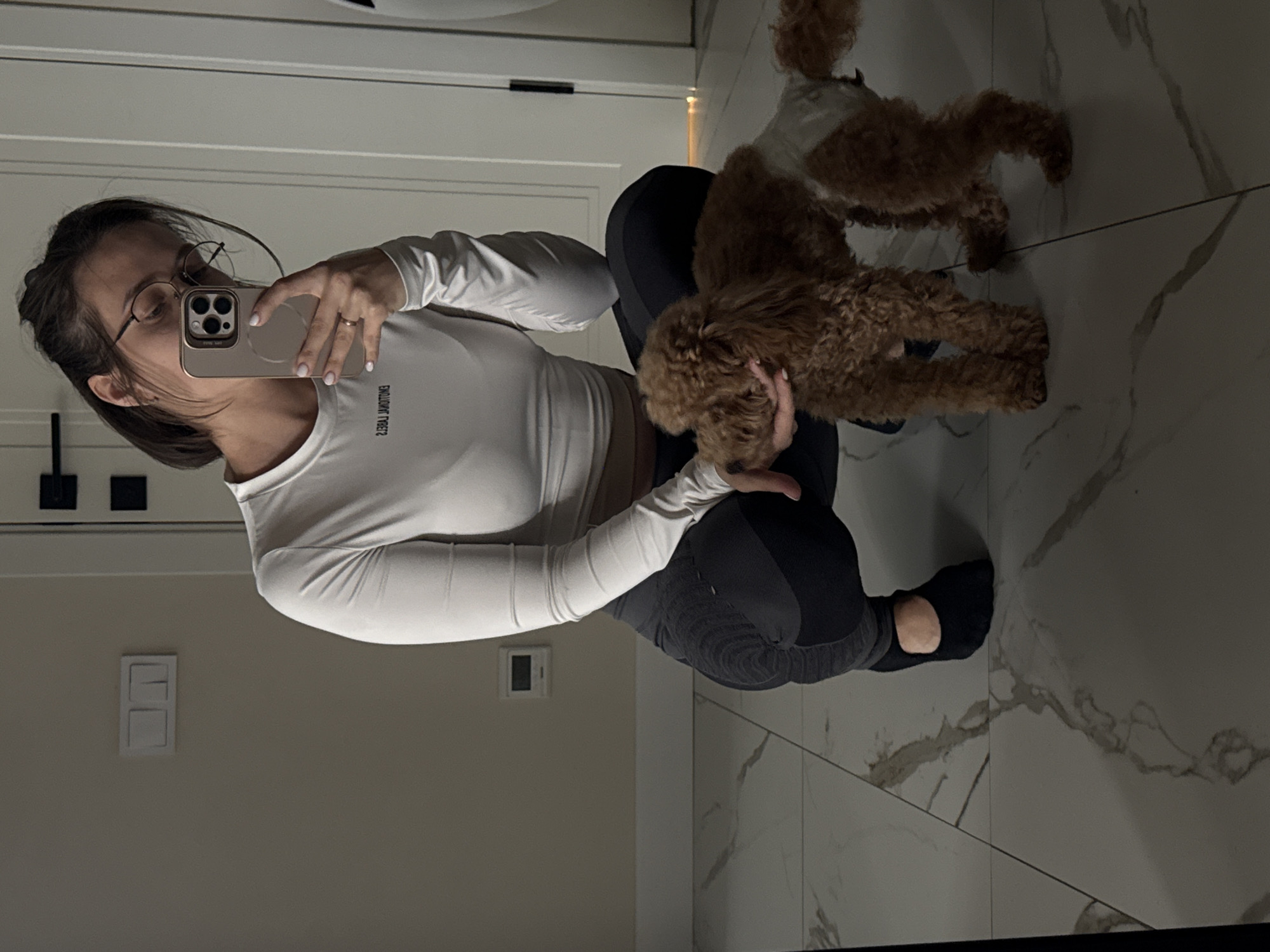 An indoor mirror selfie captures a young woman crouching on a light-colored tiled floor with two small, brown, fluffy dogs. The woman, with dark hair tied back and wearing glasses, holds a light-colored smartphone to take the picture, obscuring most of her face. She is wearing a white long-sleeve crop top and dark leggings, with a ring visible on her left ring finger. Text on her top, mirrored from the selfie, appears to read "YIELD THEORY."
One of the poodle-like dogs is in her arms, looking towards her hand, while the other stands nearby on the tiled floor. The floor features large, light-colored tiles with prominent dark, marbled patterns. The walls are a plain light color, possibly beige or off-white. On the left wall, a white wall-mounted light switch or outlet cover is visible, along with a dark, rectangular fixture higher up, possibly a handle or bracket. The overall lighting is soft and ambient, suggesting an indoor setting without direct natural light, possibly in the evening or a dimly lit room. The scene conveys a casual and affectionate moment between the woman and her pets.