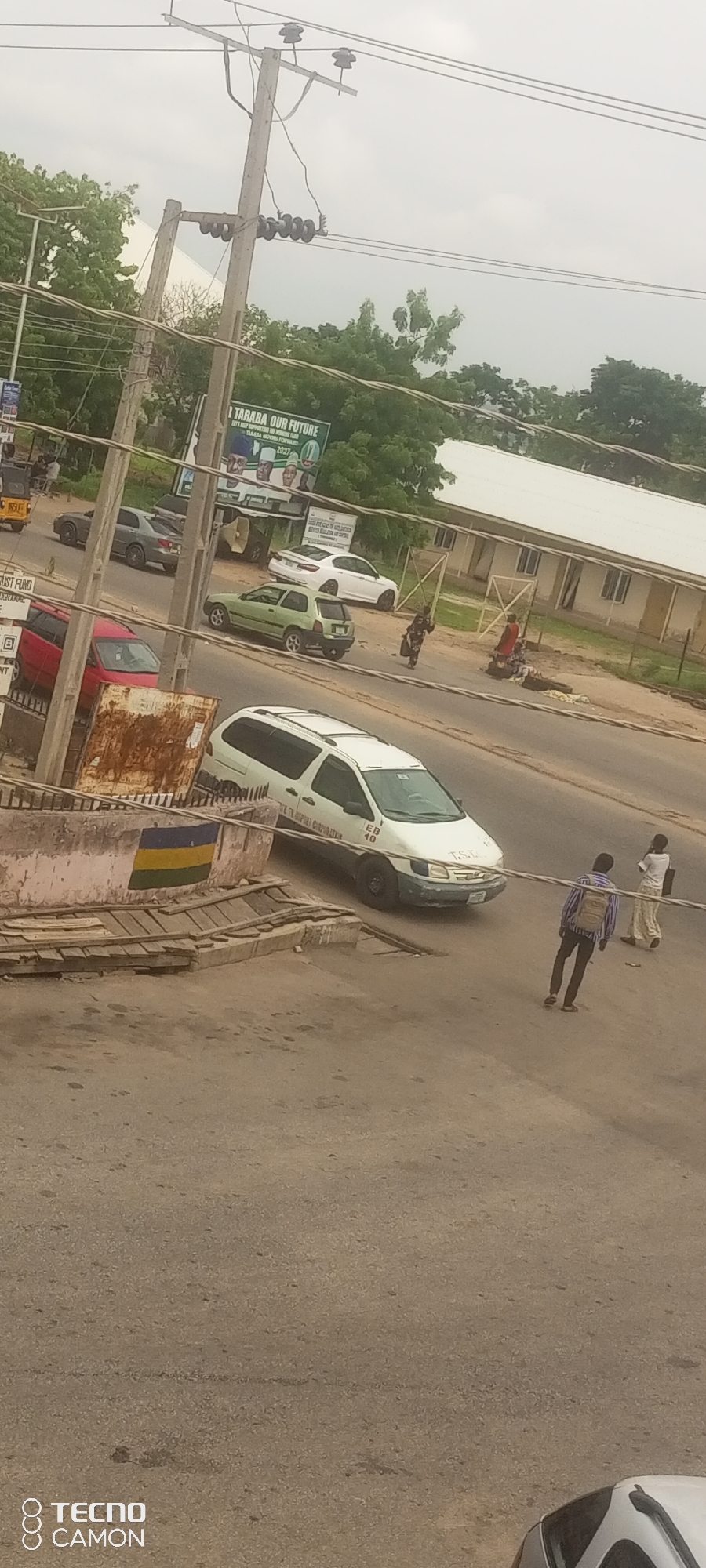 The image depicts a street scene in Jalingo, Nigeria, likely during the daytime under an overcast sky. A paved road runs diagonally across the frame, with a sidewalk and surrounding vegetation visible.
In the foreground, a white minivan is parked on the right side of the road. The minivan has "EB 40" and "T.S.L." written on its side. To the left of the minivan, a concrete structure with a flag painted on it, featuring yellow, green, and blue stripes, is present. Wooden planks are arranged on top of this structure.
Further down the road, several other vehicles are present, including a green hatchback, a grey sedan, and a white sedan. Two individuals are walking on the road, one wearing a striped shirt and backpack, and another in a red garment. Another person is seen walking on the sidewalk in the middle ground.
In the background, a large billboard with text and images of people is visible. Utility poles with wires run across the top of the frame, and lush green trees line the sides of the road. The overall environment suggests an urban or semi-urban setting.