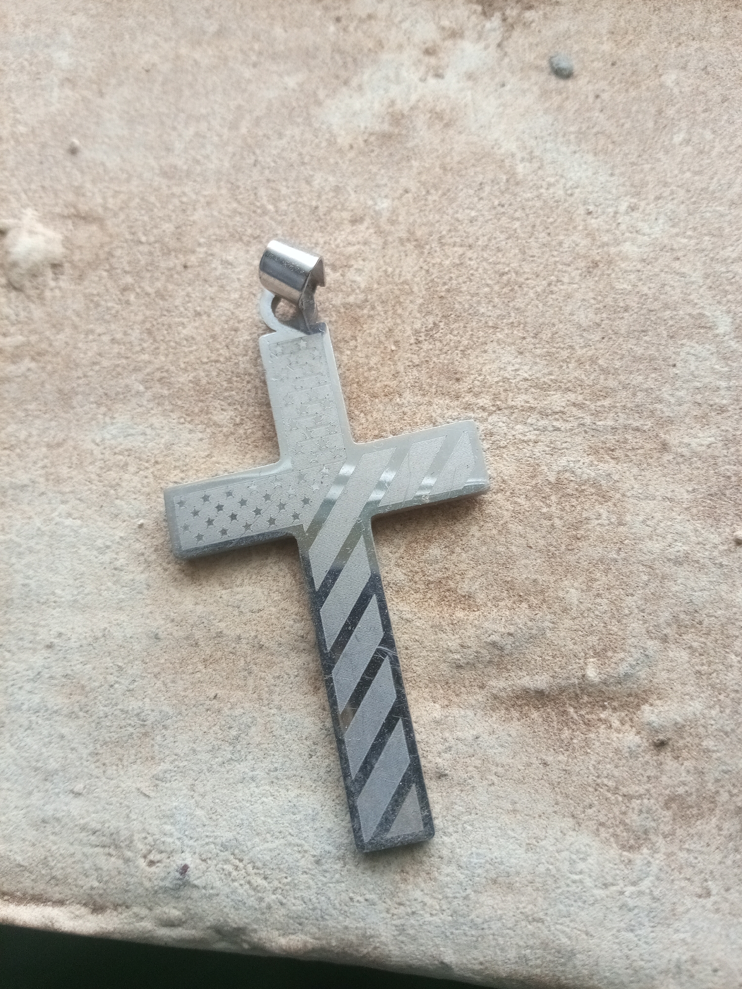 A silver cross pendant lies on a rough, sandy-colored surface, possibly concrete or stone. The cross has a textured design. The top horizontal arm of the cross is adorned with a pattern of small stars, reminiscent of the American flag. The bottom vertical arm features a striped pattern with alternating dark and light gray diagonal lines. A small, loop-like attachment for a chain is visible at the top of the pendant. The background surface is uneven and appears weathered, with some small debris scattered across it. The lighting suggests it is outdoors, possibly under natural light.