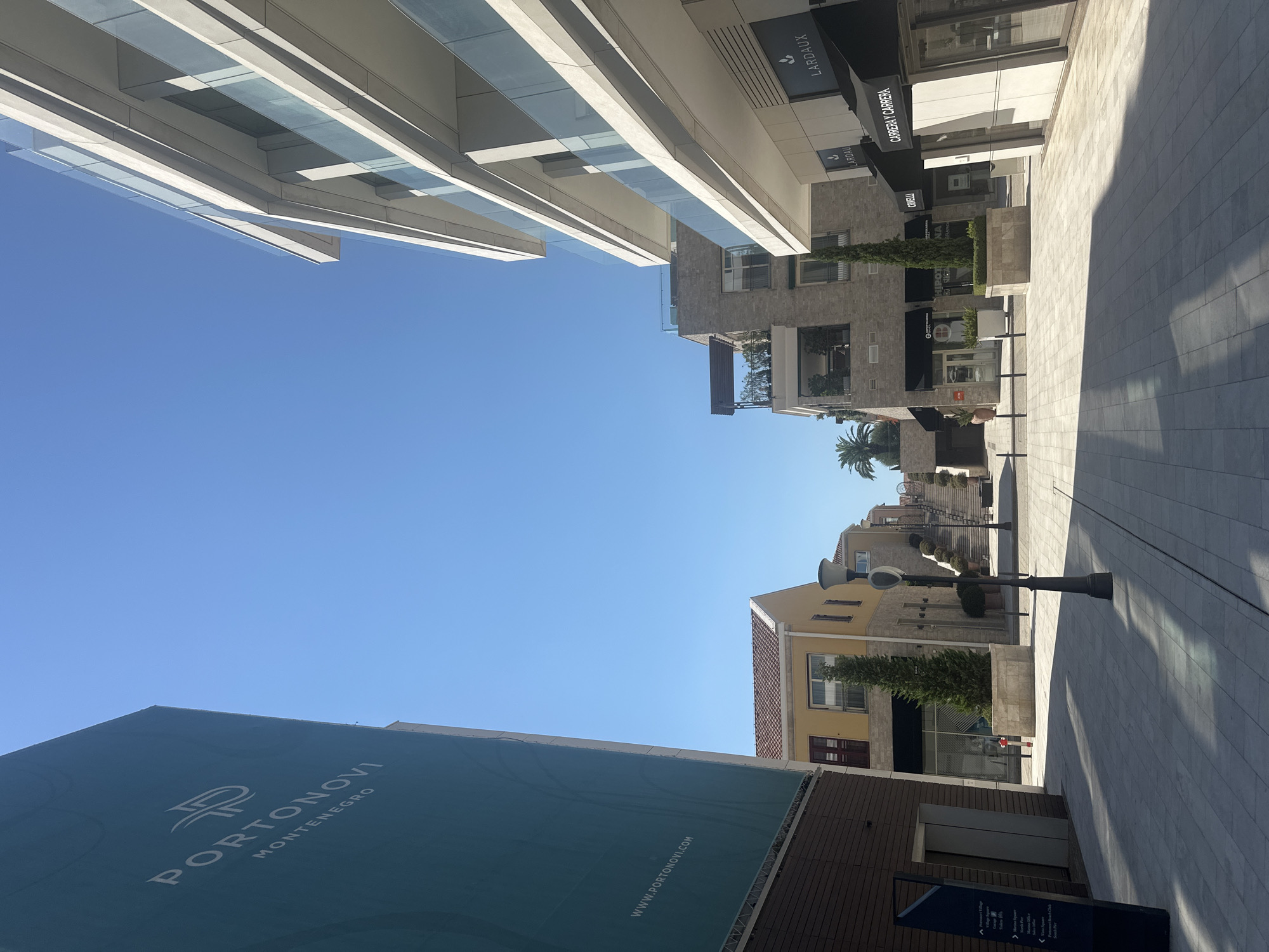 A sun-drenched, modern pedestrian street scene is captured in Kumbor, Montenegro, under a clear blue sky. To the left, a large teal banner with white lettering for "PORTONOVI MONTENEGRO" dominates the view, with "WWW.PORTONOVUI.COM" visible at its base. Adjacent to the banner is a dark brown brick-clad building.
The street itself is paved with light-colored, square tiles, casting long, sharp shadows from the buildings and a prominent black lamppost in the foreground. On the right side of the street, a contemporary white building with multiple levels features balconies with glass railings. Below, shopfronts with black awnings are visible, including one with "LARDAUX" signage and another with "CARRERA Y CARRERA."
Further down the street, a distinct yellow building with a terracotta-tiled roof stands, featuring dark red shutters on a window. A slender, conical evergreen tree is planted in a stone planter in front of it. Beyond this, the street slopes upwards, leading to a staircase flanked by neatly trimmed bushes and more lampposts, with glimpses of other buildings and palm trees in the distance.
The environment appears to be a resort or luxury development, characterized by its clean lines, well-maintained landscaping, and modern architecture. No people are actively present in the frame, contributing to a sense of stillness and quiet.