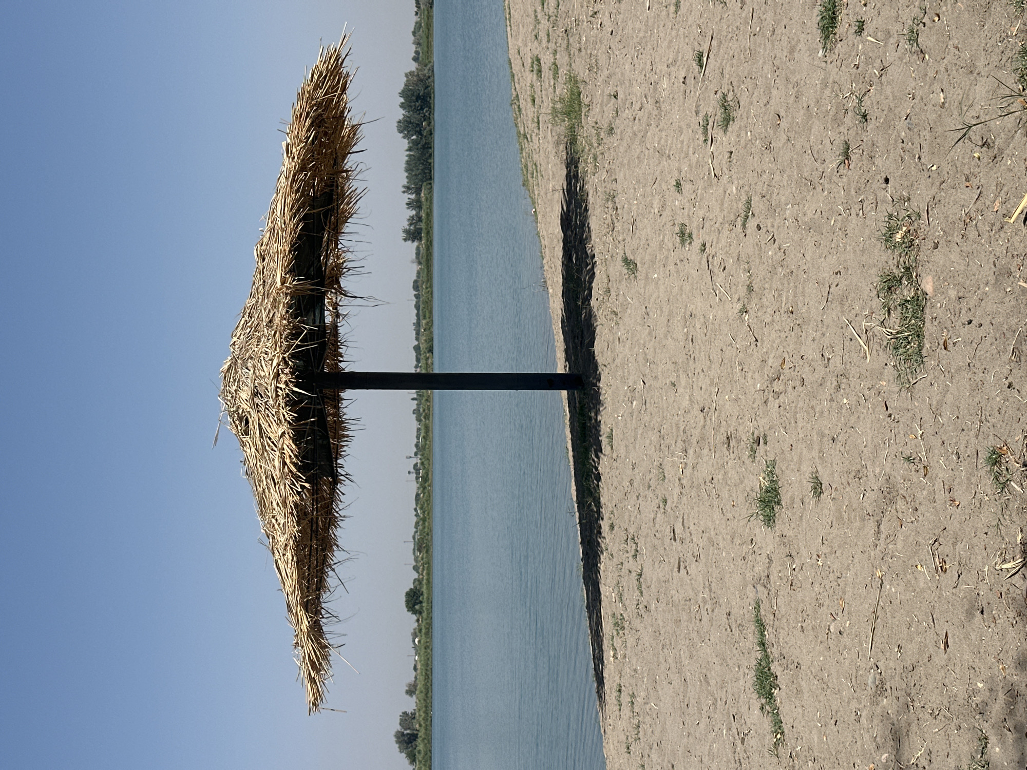 A lone thatched umbrella stands in the sand on the shore of a lake, casting a shadow. The lake is calm and reflects the clear blue sky. Across the water, a line of trees and buildings can be seen on the horizon. The sand is dry and sparse with patches of green grass. The image appears to be taken in the city of Dehkanabad, Uzbekistan.