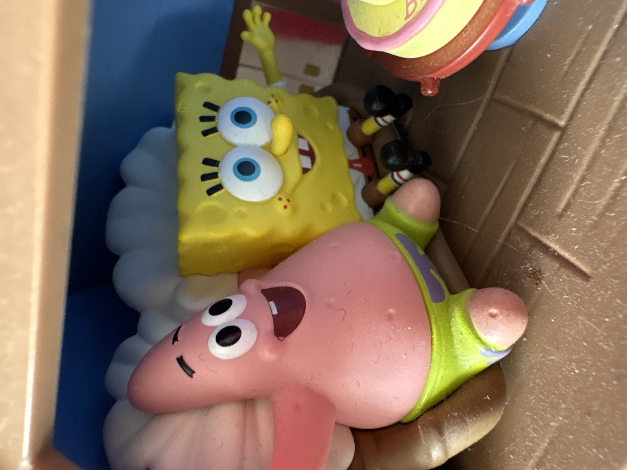 The image displays two cartoon characters, Spongebob Squarepants and Patrick Star, from the animated television series *Spongebob Squarepants*. Spongebob is depicted as a yellow, porous square with large blue eyes and a wide smile. He is wearing white shorts and black shoes with red and white striped socks. Patrick Star is a pink, star-shaped character with black eyes and a similar wide grin. He is wearing green shorts with purple trim.
They appear to be part of a toy set, possibly a playset, with a brown plastic textured floor and a blue background. Spongebob is positioned behind Patrick, with his body partially obscured by what looks like a white, cloud-like molded plastic element. Patrick is in the foreground, lying on his back, with his arms and legs spread. Spongebob has one hand raised, as if waving.
In the upper right corner of the image, a portion of another toy is visible. It is round and yellow with pink and red accents, and some faint purple lettering that might be part of the word "BEST".
The lighting suggests an indoor setting, likely with artificial light. There are no clear indicators of time of day or weather. The overall scene is playful and reminiscent of the cartoon's lighthearted tone. The toys appear to be plastic and are rendered in bright, vibrant colors typical of children's toys. The surface they are on has a grid-like texture. There is some dust or lint visible on the brown surface, suggesting it may not be perfectly clean.