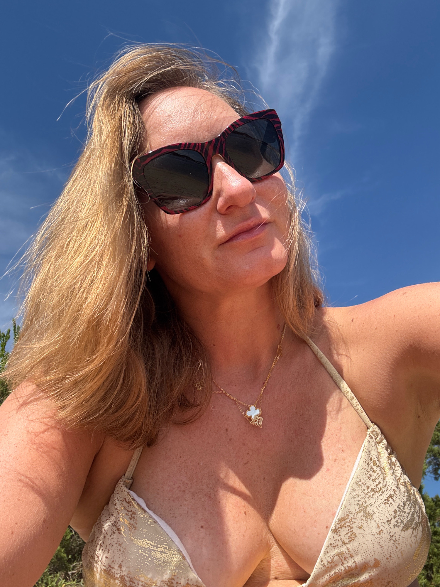 A woman is taking a selfie outdoors, with a bright blue sky and some wispy clouds behind her. She has blonde hair and is wearing sunglasses and a gold bikini. She is also wearing a delicate gold necklace with a white clover-shaped pendant. The sun is shining brightly, casting a shadow on her chest. The background suggests a natural setting, possibly near the coast, given the greenery visible at the bottom of the frame. There are no visible landmarks to pinpoint the exact location within Lu Palau/Palau, Italy.