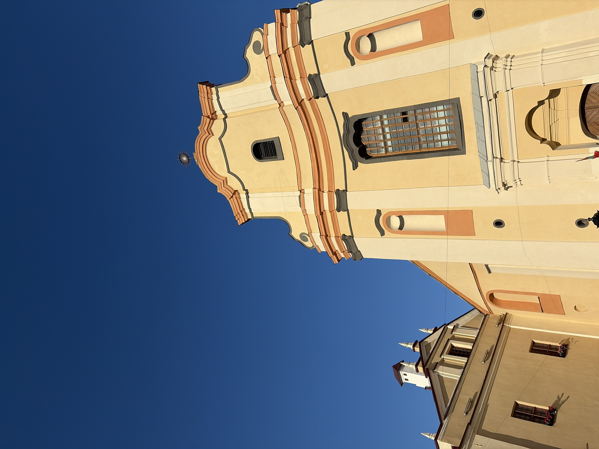 The image shows the exterior of the Holy Spirit Cathedral in Minsk, Belarus, against a clear blue sky. The cathedral is characterized by its yellow facade with decorative orange and gray accents. A prominent feature is a tall, arched window with a wooden frame. To the left, another building with a pitched roof and dormer windows is visible. The image is taken from a low angle, emphasizing the architecture of the buildings. No people are present in the frame.