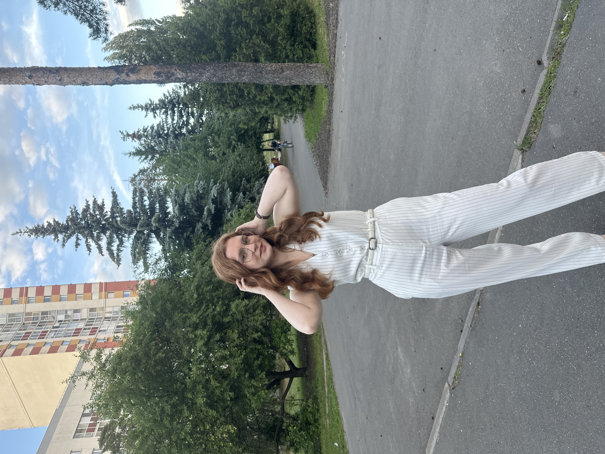 The image features a young woman with long, wavy red hair and eyeglasses, standing on a paved path in an outdoor park setting. She is wearing a white pinstriped jumpsuit with a white belt, and has a watch on her left wrist. Her hands are raised, resting gently near her head, and she is looking towards the viewer with a calm expression.
The background is a mix of natural elements and urban architecture. Dominating the scene are numerous lush green trees, including tall evergreens and deciduous trees, with a large tree trunk visible prominently on the left side of the path. The sky above is blue with scattered white clouds. To the far left, a tall, multi-story building with a distinctive facade featuring rectangular panels of orange, yellow, and white or grey is visible.
Further down the paved path, two distant figures can be seen, suggesting other people are present in the park. The ground is asphalt, showing some cracks with grass growing through them along the edges of the path. The scene depicts a pleasant daytime moment in a green urban environment, consistent with a park in a city like Kazan, Russia. There is no visible text in the image.