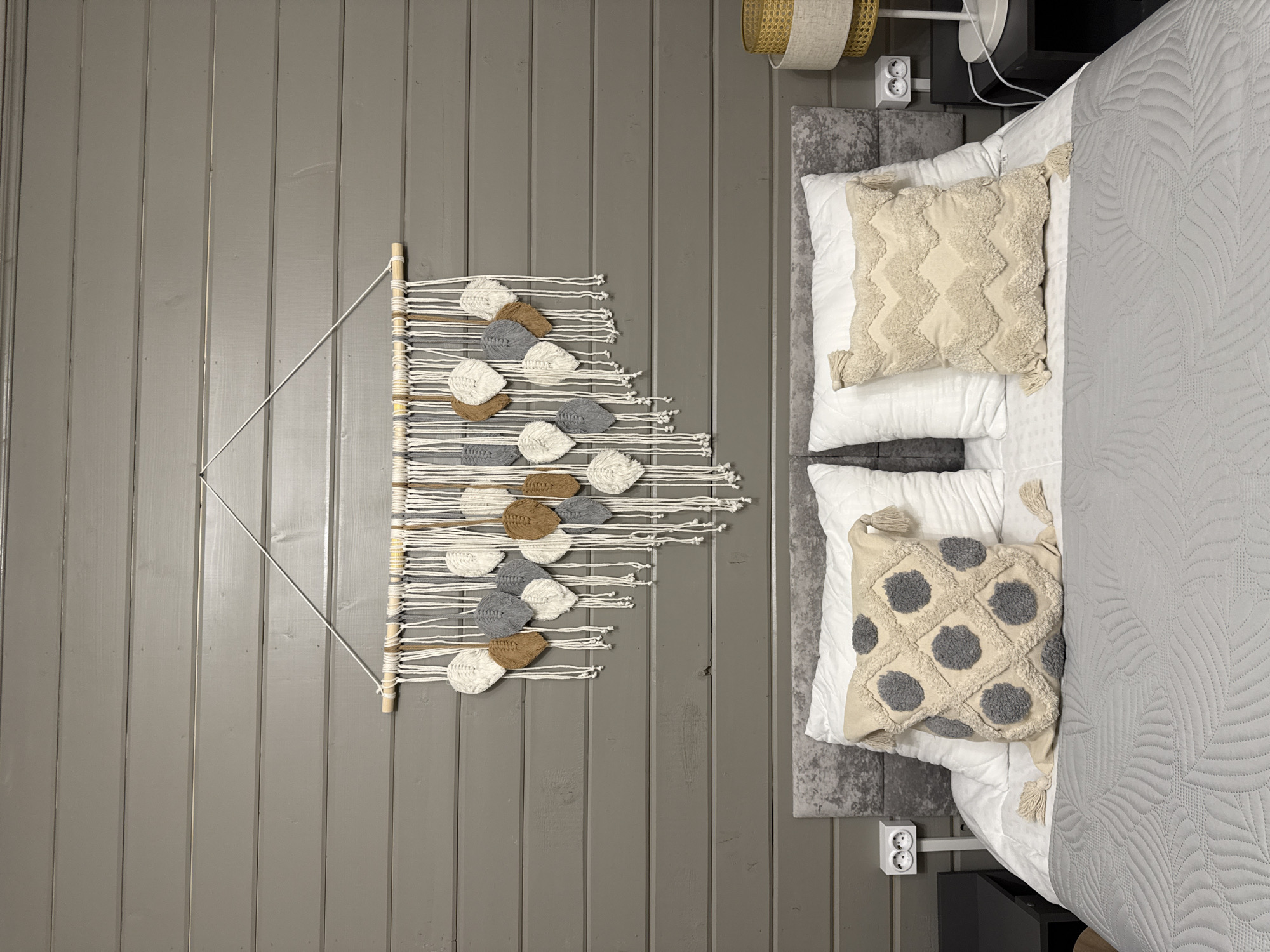 An overhead shot of a bedroom interior, possibly in Buzhaninovo, Russia, reveals a gray-painted wooden wall on the left. Mounted on this wall is a macramé wall hanging. The wall hanging consists of a wooden dowel from which several white, gray, and brown leaf-shaped decorations hang, attached by horizontal strings. To the right, a portion of a bed is visible. The bed is dressed in white linens and gray bedding with a leafy pattern. Two decorative pillows rest on the bed: one with a geometric textured design in cream, and another with gray pom-poms. A gray rug is under the bed. On the right side of the bed is a black nightstand. A basket and electrical outlet are present near the head of the bed. The scene appears to be a still life, presenting a neat and styled domestic environment.