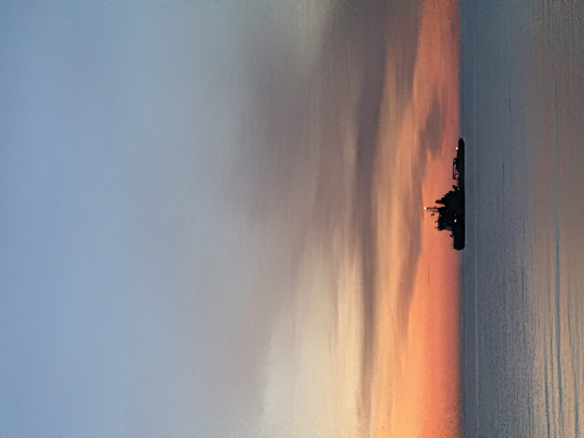 The image shows a cargo ship silhouetted against a sunset sky over the Baltic Sea. The sky displays a gradient of colors, transitioning from pale blue at the top to vibrant orange and pink hues near the horizon. The sea is calm with gentle ripples reflecting the sky's colors. The ship, a prominent dark silhouette, is positioned centrally in the frame. The scene appears to be captured from a distance, possibly from the coast or another vessel. The overall atmosphere is serene and evocative of twilight. The location is Tallinn, Estonia, and the time of day is sunset.
