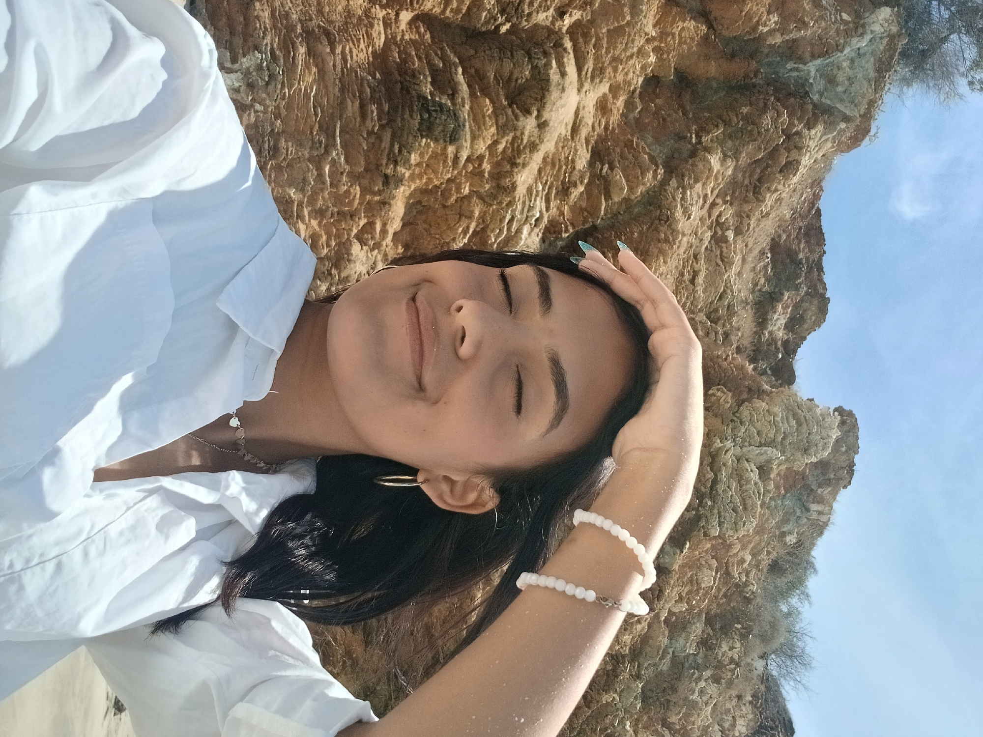This vertically oriented photo captures a young woman in a serene, relaxed pose outdoors, likely in Juan Griego, Venezuela. The woman is positioned with her head tilted, eyes closed, and a gentle smile, suggesting a moment of peace or contentment. Her dark hair falls around her shoulders. She wears a white collared shirt, open at the neck, revealing a silver necklace with multiple small heart charms. In her ear, a gold hoop earring is visible. Her right arm is raised, with her hand near her head, and she wears two white beaded bracelets on her wrist. Her fingernails are painted with a light blue or teal polish, possibly with a glitter effect. A fine layer of sand or dust is visible on her arm.
The background consists primarily of a rugged, textured cliff face in shades of brown and tan, indicating a natural, rocky environment. Above and to the right of the woman's head, a clear, bright blue sky with a faint wispy cloud is visible. The strong, direct lighting suggests it is daytime, likely mid-morning or afternoon, casting some shadows on her shirt. The overall mood is one of calm and natural beauty, consistent with a coastal or natural landmark setting in Venezuela. There is no visible text in the image.