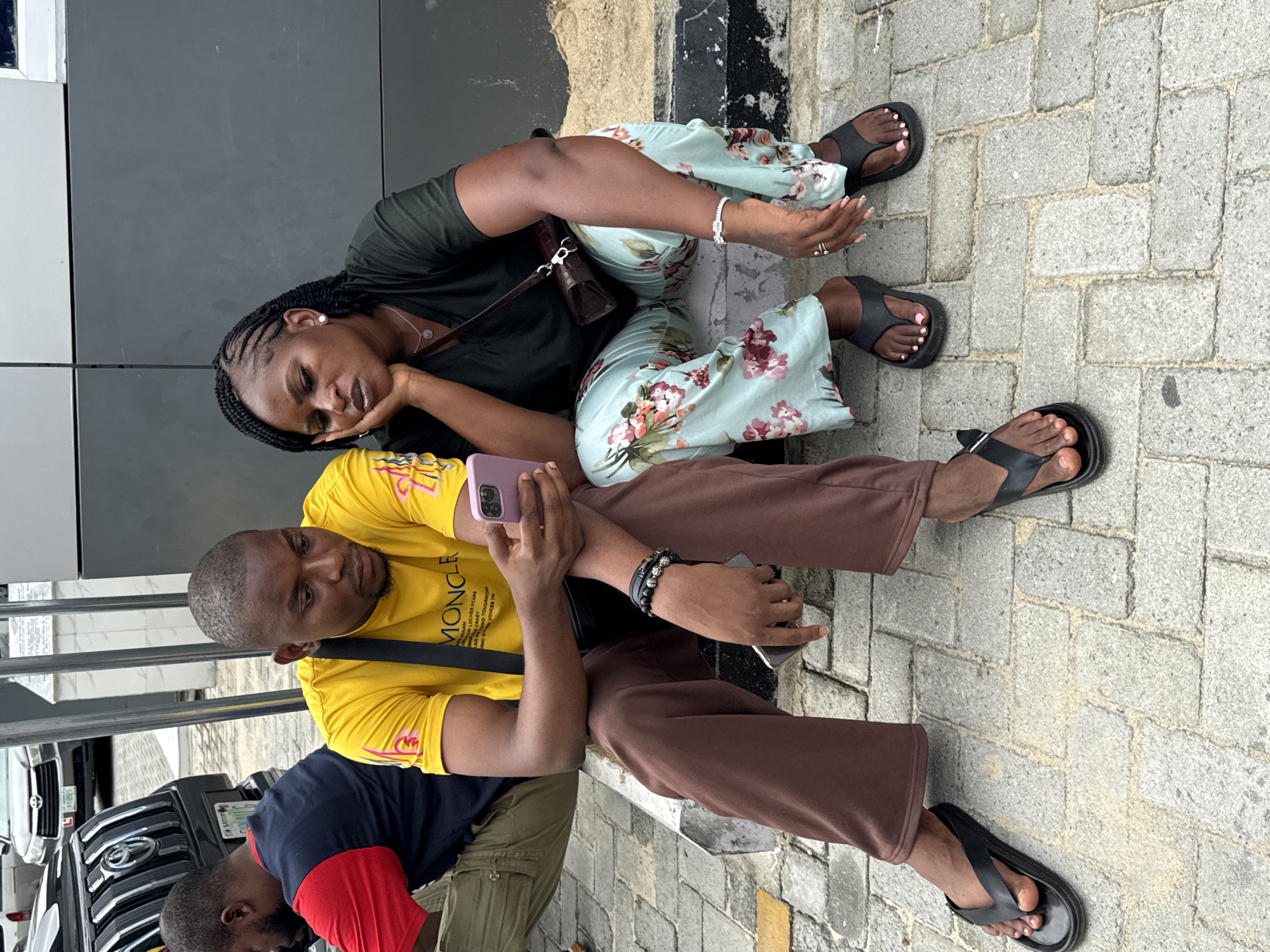 Two individuals are seated on the ground in an urban setting in Ojomu, Nigeria. The woman, who is on the left, wears a dark green top and floral print pants, with her head resting on her hand. The man wears a yellow shirt and brown pants and is holding a mobile phone. Both are wearing sandals. The background shows a dark gray wall and a brick pavement. A car is partially visible in the lower left corner.