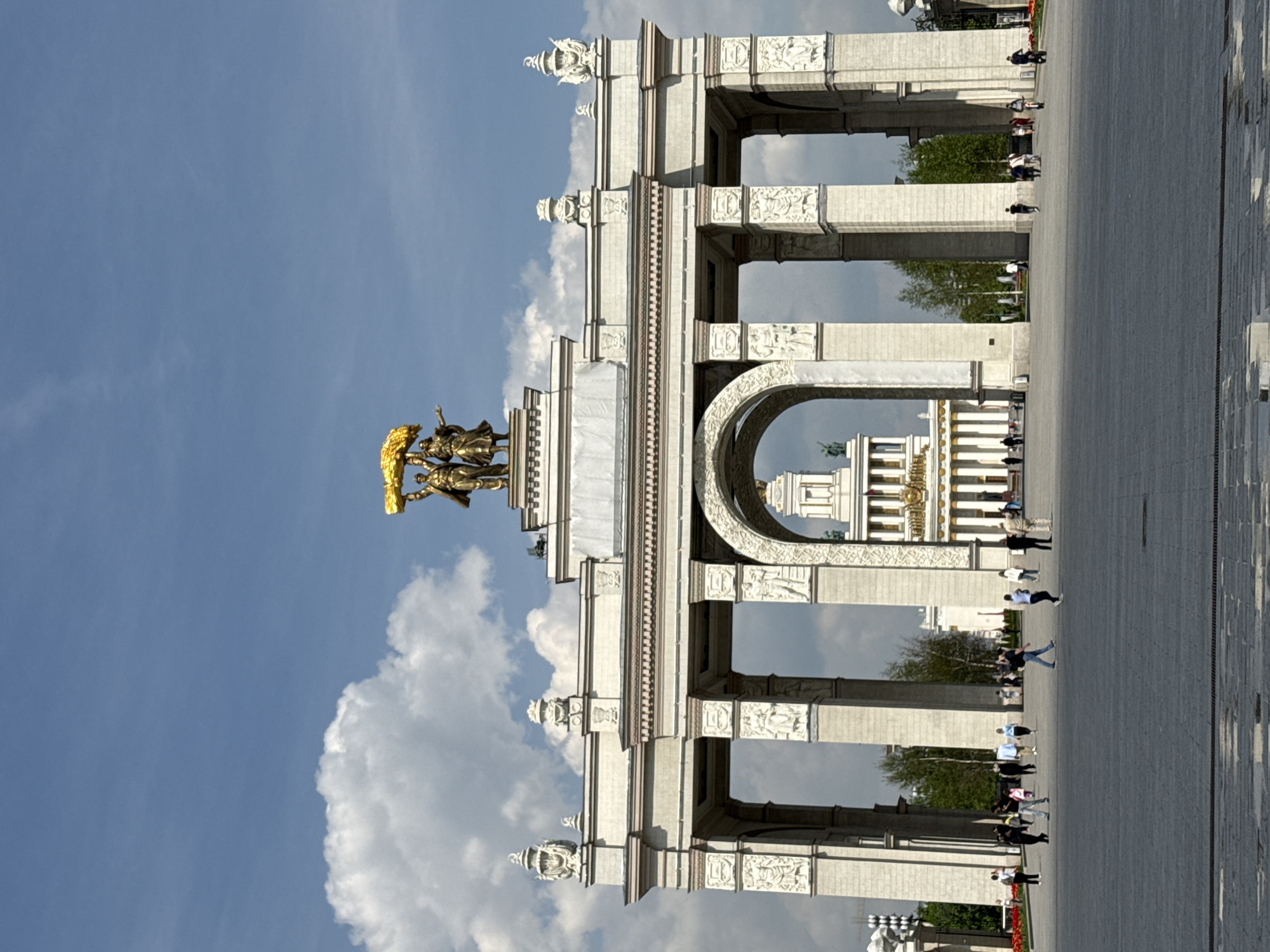 This is a photograph of the Central Arch of the main entrance to the VDNKh Exhibition Park in Moscow, Russia. The arch is made of white stone and features ornate decorations and sculptures. At the top is a golden statue of a worker and a farmer. The sky is blue with white clouds. People are walking through the arch and across the paved square. In the distance, other buildings of the exhibition park can be seen through the archway.