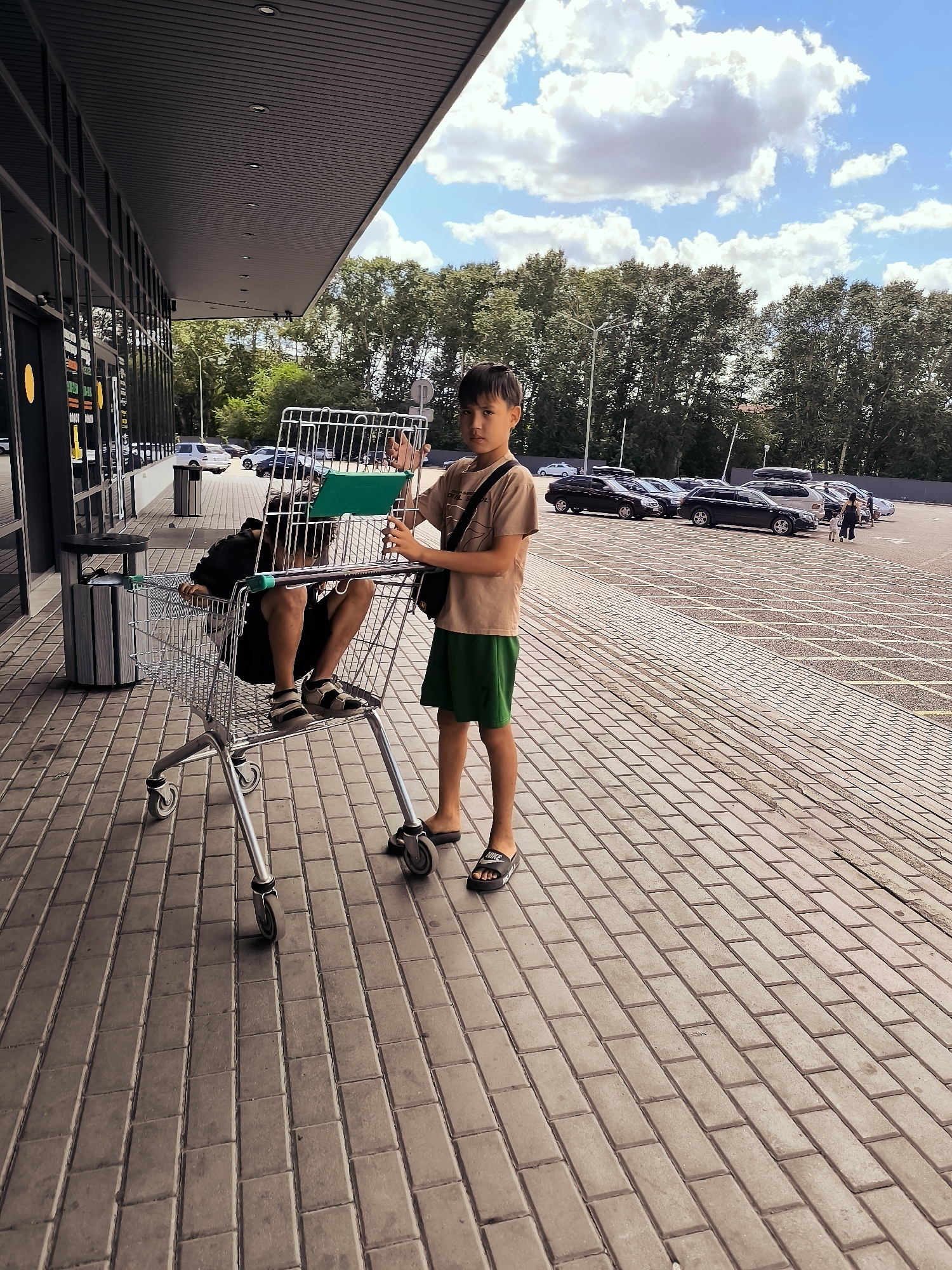 In Qarağandı city administration, Kazakhstan, a young boy stands next to a shopping cart on a paved outdoor area. The boy is wearing a tan t-shirt and green shorts, with dark hair and sandals. He is holding onto the shopping cart. Inside the shopping cart, another person, wearing a black shirt and dark shorts, is sitting down. The shopping cart is made of metal and has wheels. In the background, there is a building with a dark roof and glass windows, trees, and a parking lot with several cars. The sky is blue with white clouds. The ground is covered in rectangular paving stones.
