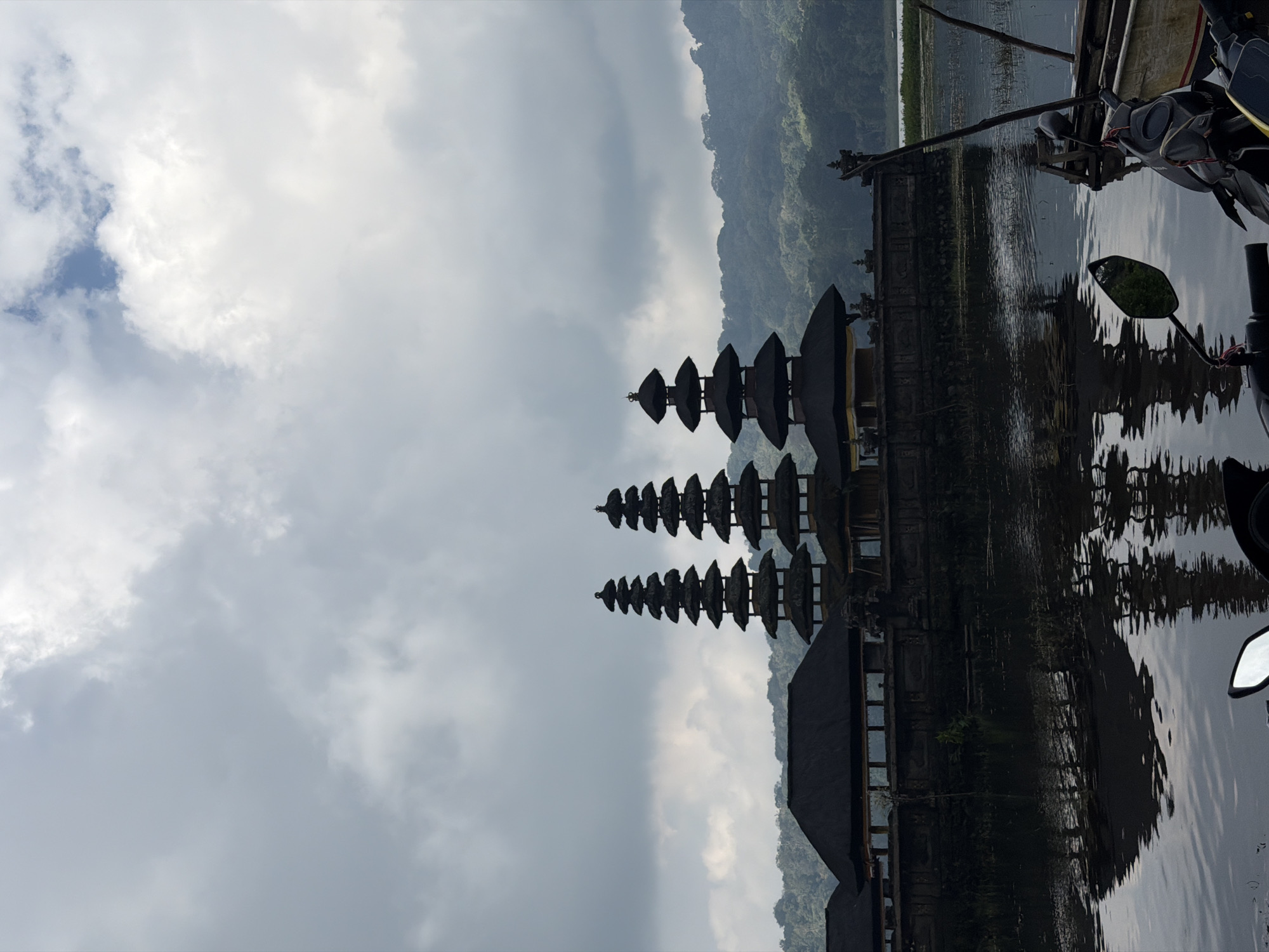 This is a photograph of the Pura Ulun Danu Bratan temple complex in Banjar, Indonesia. The temple is situated on the edge of a lake, with its distinctive tiered pagodas reflected in the water. In the foreground, two motorcycles are parked, with one of their mirrors visible in the lower right corner of the frame. The sky is overcast with dramatic clouds, and the surrounding hills are covered in lush green vegetation. The overall mood of the image is serene and atmospheric, conveying a sense of peace and natural beauty.