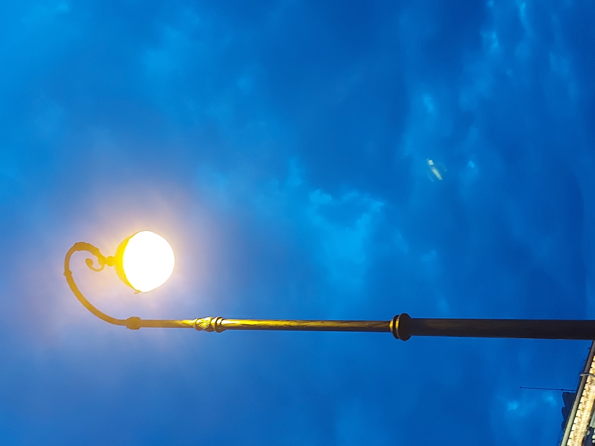 The image shows a street lamp illuminated against a dark blue sky with scattered clouds, likely taken at dusk or night. The lamp has a curved, ornate arm supporting a spherical light. The lamp post appears metallic, with some golden-toned sections. Part of a building's rooftop is visible in the bottom right corner. The location is most likely Saint Petersburg, Russia based on the architecture of the lamppost.