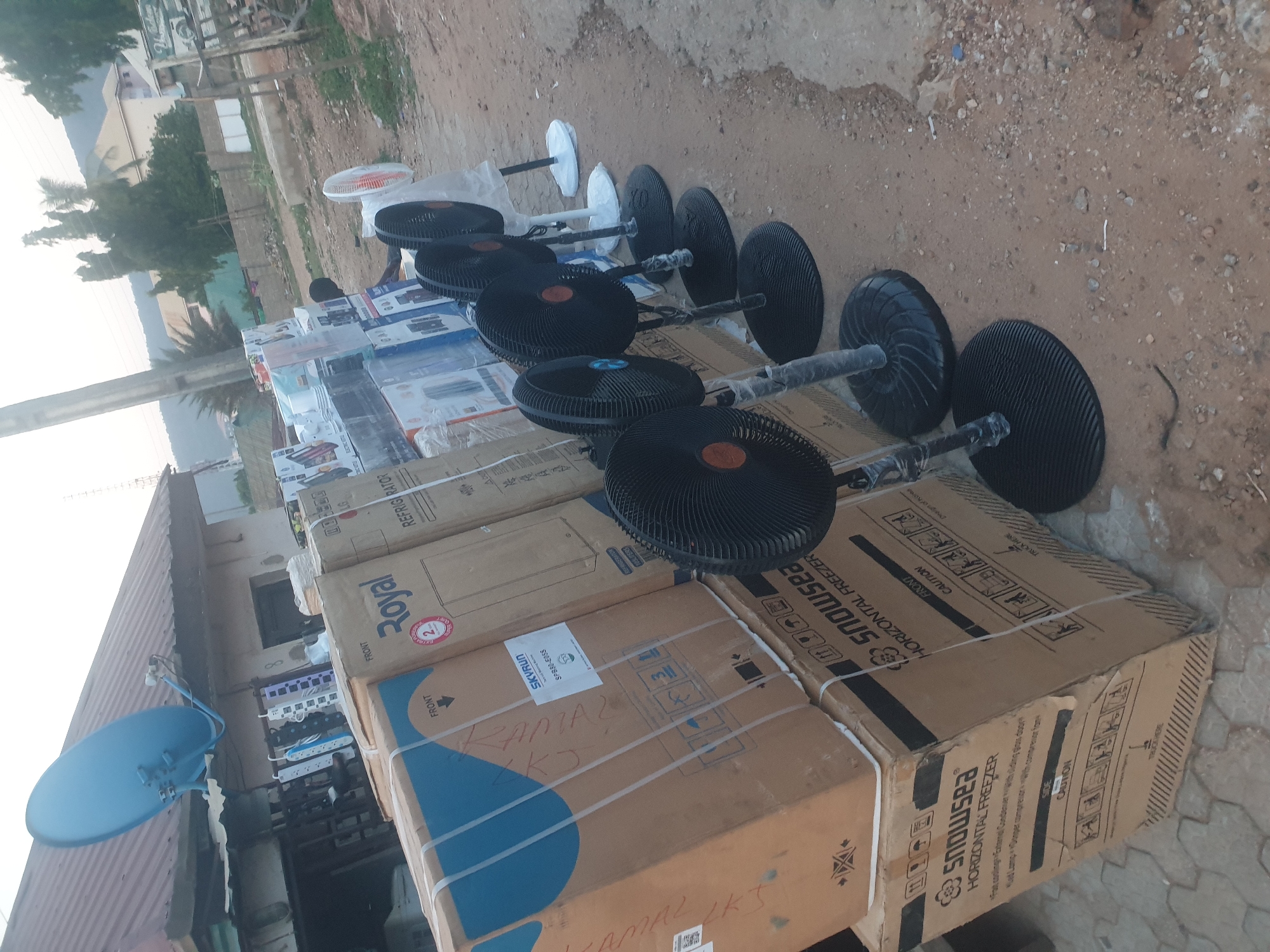 Here is a detailed description of the image:
**Location:** Adankolo, Nigeria.
**Scene:** An outdoor setting, likely a street or open area near a shop or marketplace.
**Objects:**
* Several cardboard boxes are stacked.
* Some boxes are labeled "Snowsea Horizontal Freezer."
* One box is labeled "Royal."
* One box is labeled "LG Refrigerator".
* One box has the name "KAMAL LKS" written on it.
* The boxes are secured with white plastic straps.
* Several floor fans in their packaging or with their bases protected.
* A blue satellite dish is visible, mounted on a building.
* A line of electrical power strips are visible on the side of a building.
**Environment:**
* The ground is dusty or sandy.
* There are trees in the background.
* Buildings of varied construction are visible.
* The lighting suggests it may be late afternoon or early morning.
**Overall Impression:** The scene depicts goods, specifically appliances and floor fans, likely for sale or recently delivered in the city of Adankolo, Nigeria.