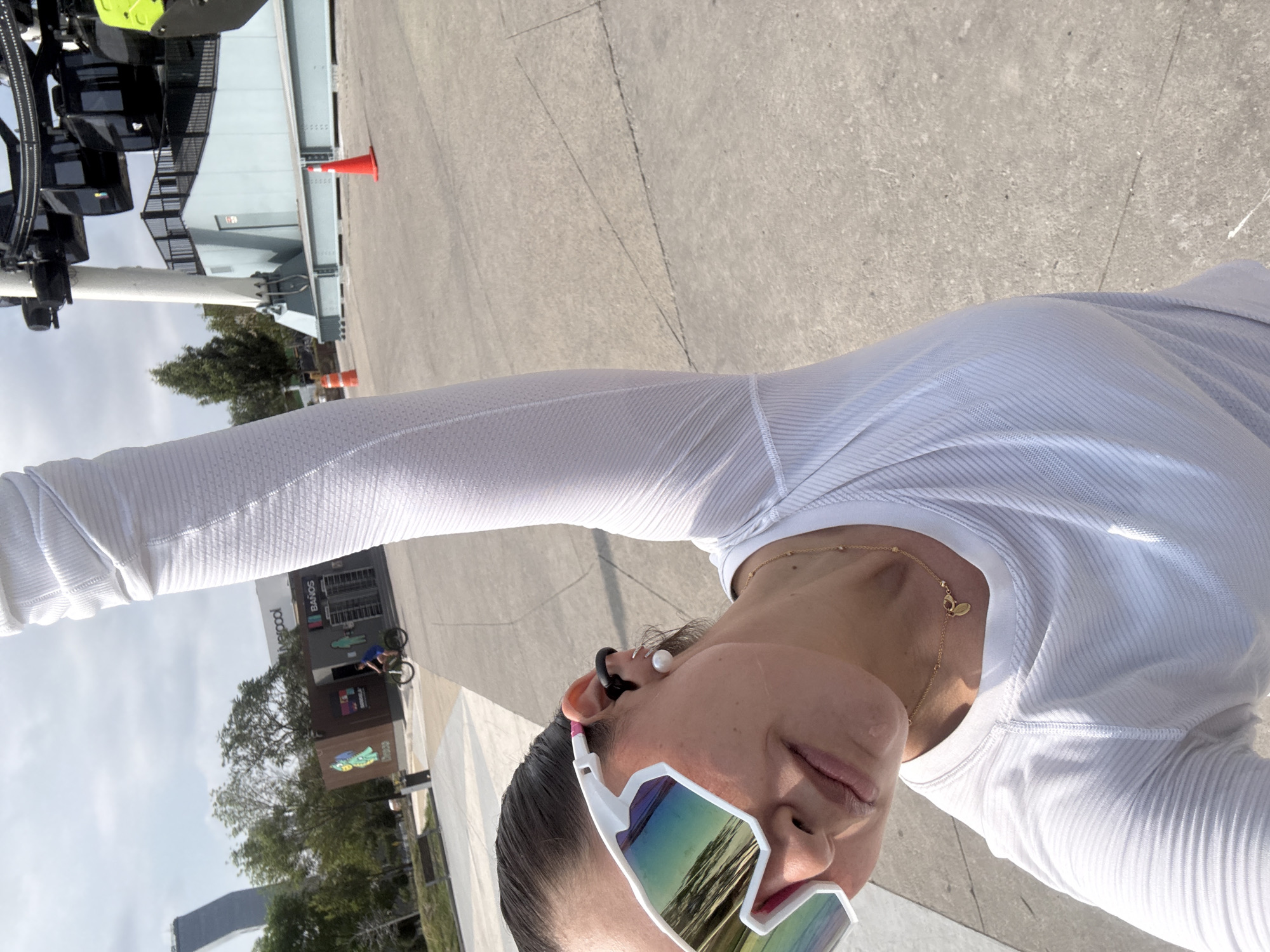A woman is taking a selfie outdoors in Puebla, Mexico. She is wearing a white long-sleeved shirt, white sunglasses, and has an earpiece in her left ear. In the background, there is a gray concrete surface, and on the left side of the image, a tall metallic structure is visible along with trees and the corner of a building with the word "Liverpool" on it. A person on a bicycle is visible next to the building. There is also an orange cone in the background.
