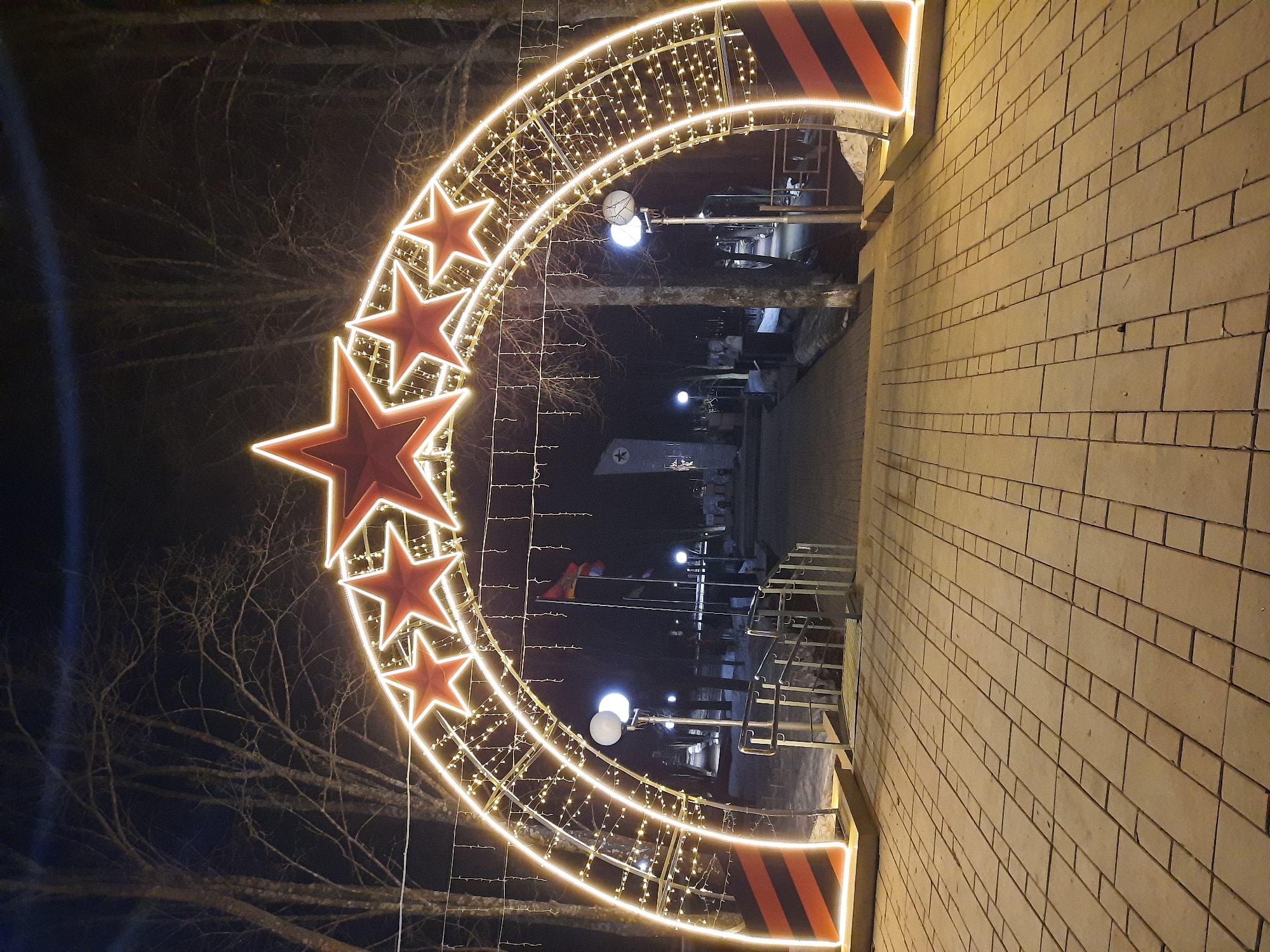 The image shows a nighttime view of an illuminated archway in Krasnogorskoye, Russia. The arch is decorated with multiple red stars outlined in white lights, and further adorned with strings of small white lights. The archway frames a walkway, visible in the background, which leads into a dimly lit area. There are lamp posts lining the walkway. To the right of the arch is a brick wall. Bare trees are visible to the left of the archway. The scene appears to be part of an outdoor public space.