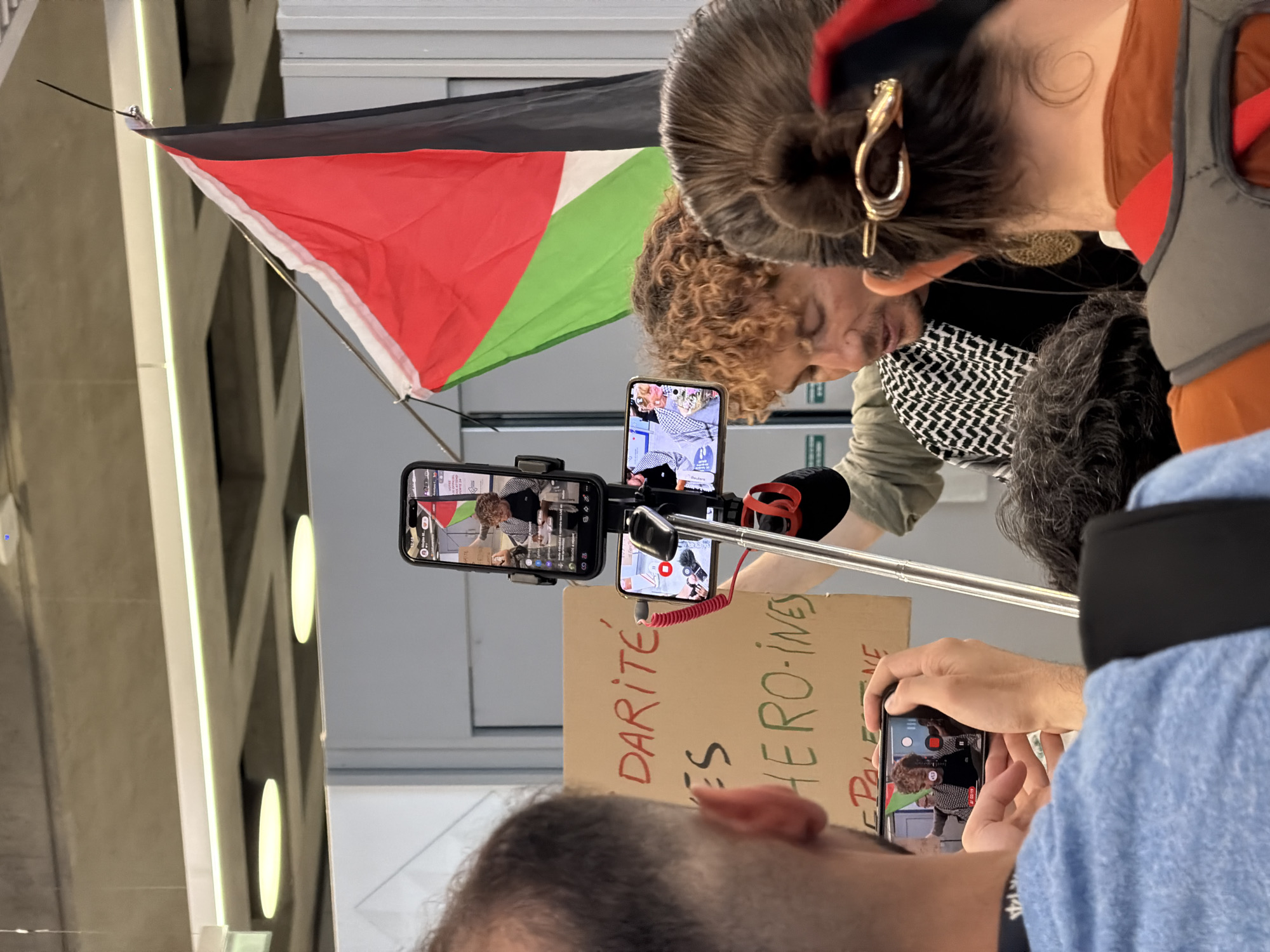 The image shows a demonstration or protest, likely in an indoor setting, with a flag of Palestine visible in the upper left corner. Several people are present, and some are using smartphones to record or stream the event. One person has two phones mounted on a rig with a microphone, pointed towards a cardboard sign that reads "DARITE ES HERO-INES", possibly referring to Palestinian heroines. Another person is taking a picture with a smartphone. The ceiling structure suggests a modern building. The location is Mauregard, France.