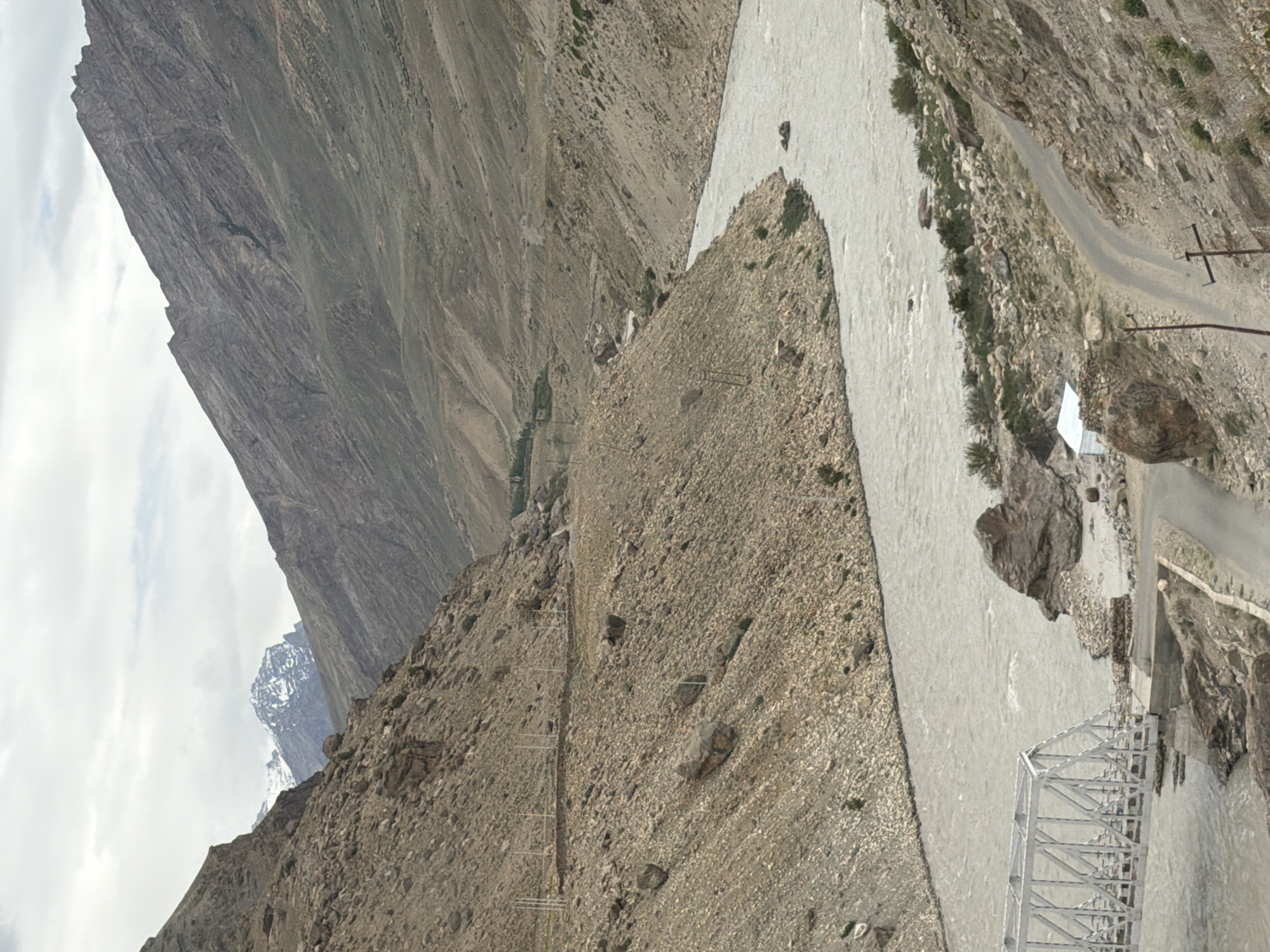 The image depicts a wide, milky-grey river flowing through a valley in the Himalayas. The river is surrounded by steep, rocky mountains with sparse vegetation. A white truss bridge spans the river on the left side of the image. A narrow, winding road is visible on the right side of the valley, adjacent to the river. In the background, snow-capped mountains can be seen against a cloudy sky. The scene suggests a remote, mountainous location, likely in the Zanskar region of Ladakh, where Padum is located. There are no people visible in the image.