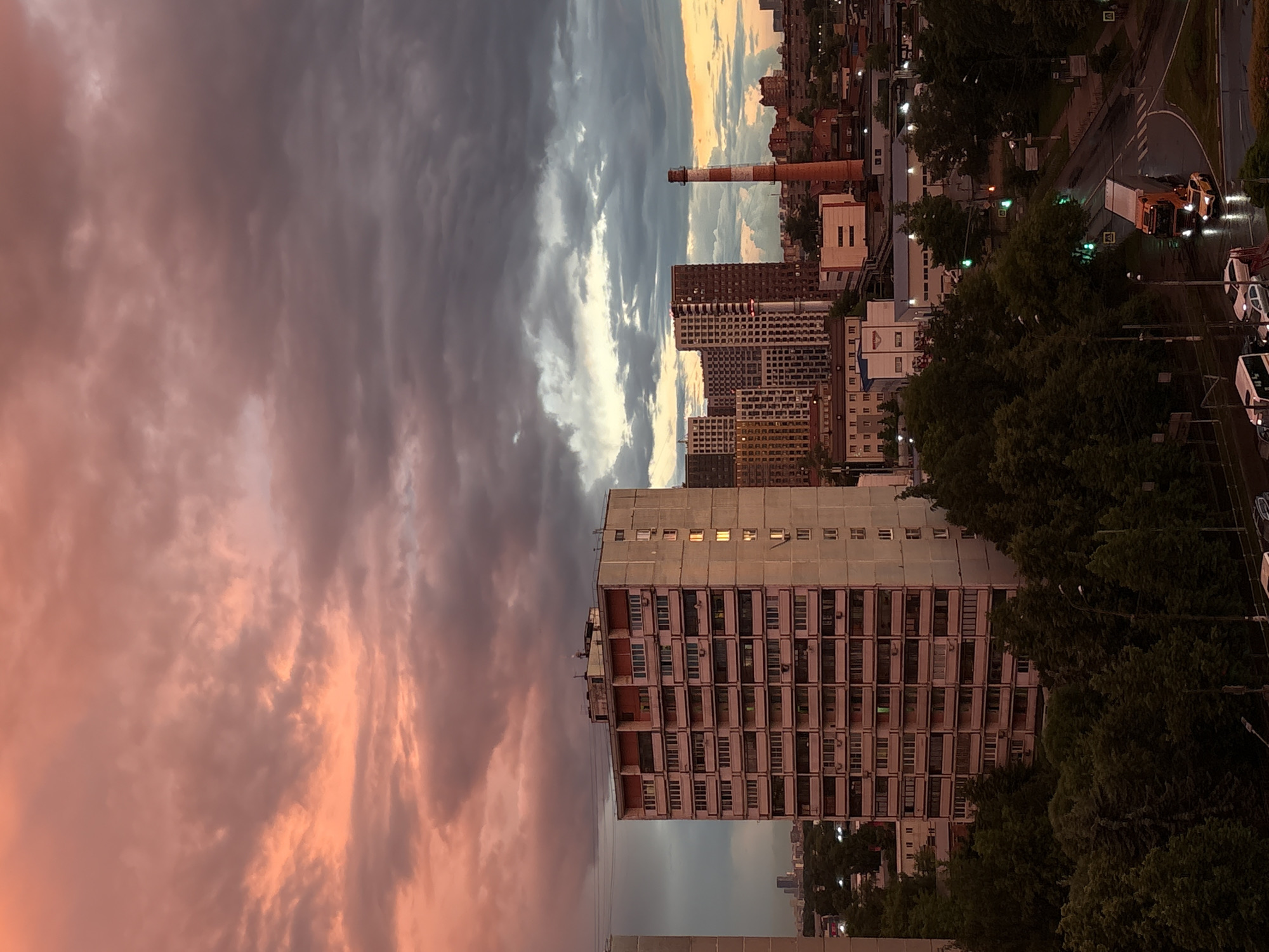 This is a vertical, medium-distance shot of an urban landscape during a dramatic sunset. The sky dominates the upper portion of the image, filled with dark, heavy clouds tinged with vibrant coral and pink hues from the setting sun.
In the foreground, a tall, multi-story apartment building with a light-colored facade and numerous balconies is the most prominent structure. Its windows reflect the sky's colors, and some appear to be lit from within. To the right of this building, a cluster of modern, high-rise buildings with varied architectural styles and colors are visible. Further to the right, a tall, red and white striped industrial chimney stands against the sky.
The middle ground features a dense canopy of dark green trees, obscuring much of the ground level. A road curves through the scene, illuminated by traffic lights and the headlights of vehicles. A large orange truck with a trailer is in motion on the wet road, with a yellow car following behind. The wet road surface suggests recent rain.
In the far distance, a hazy cityscape with faint, silhouetted buildings can be seen. The overall atmosphere is moody and evocative, characterized by the contrast between the soft, colorful sky and the stark, geometric forms of the urban architecture. The scene appears to be set in an industrial or residential area on the outskirts of a city.
