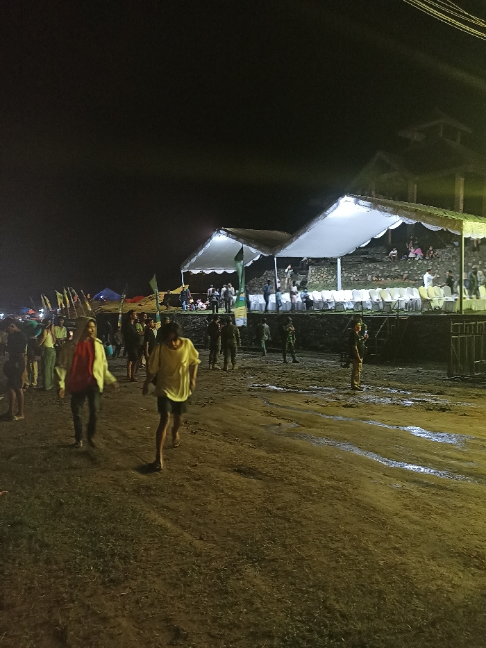 This night-time image captures an outdoor public event in Kecamatan Pujut, Indonesia. The scene is illuminated by artificial lights, contrasting with the dark sky above.
In the foreground, two individuals walk towards the viewer on an uneven dirt path, which appears wet or muddy in places, reflecting light. One person wears a bright yellow short-sleeved shirt and dark shorts, while the other is dressed in a red shirt and a light-colored jacket. Numerous other people are scattered across the middle ground, some standing and observing. Several individuals in uniform, possibly security personnel, are visible near the temporary structures.
The background features two prominent white canopies, brightly lit from beneath, suggesting stages or viewing areas. White chairs are arranged in rows in front of these canopies, indicating audience seating. Behind these structures, an elevated, terraced area, possibly made of stone or concrete, holds more attendees. To the far left, several smaller tents and numerous flags add to the festive atmosphere. A taller, multi-story building or structure is partially visible behind the canopies to the right.
The overall impression is that of a bustling outdoor gathering or festival taking place at night, with temporary setups and a significant number of participants and observers. The damp ground suggests recent rain or general moisture in the area. There is no discernible text visible in the image.