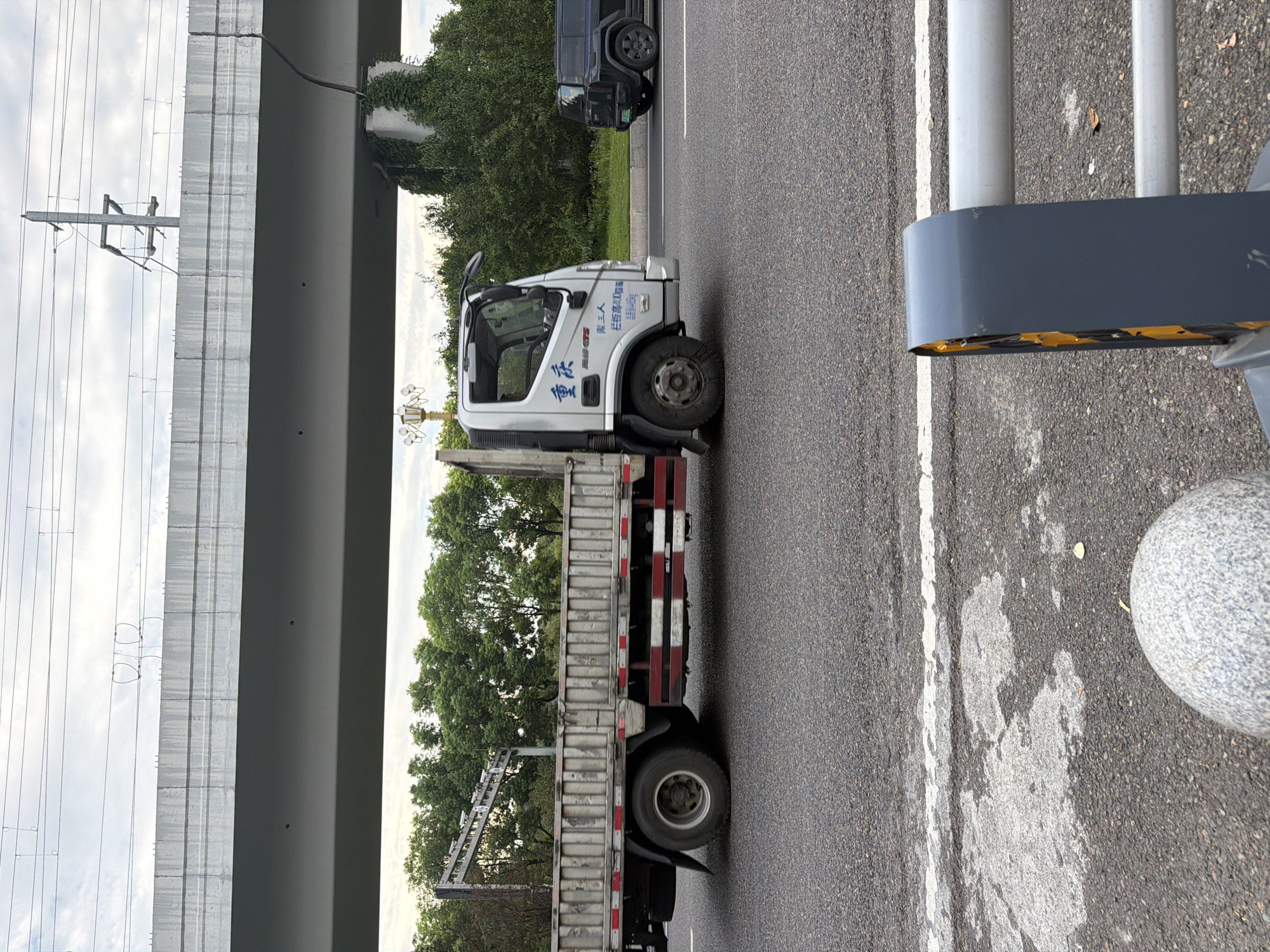 In Jiangjin, China, a silver flatbed truck with red and white reflective stripes along its sides is traveling on a paved road. The truck has Chinese characters visible on its cab, including "重庆" which translates to Chongqing. The truck is positioned on the right side of the road, with a black SUV in the lane to its right. Overhead, a large concrete overpass with visible electrical wires runs across the frame. The surrounding environment includes lush green trees and a cloudy sky. In the foreground, a white line is painted on the road, and a portion of a grey barrier with yellow reflective tape is visible.
