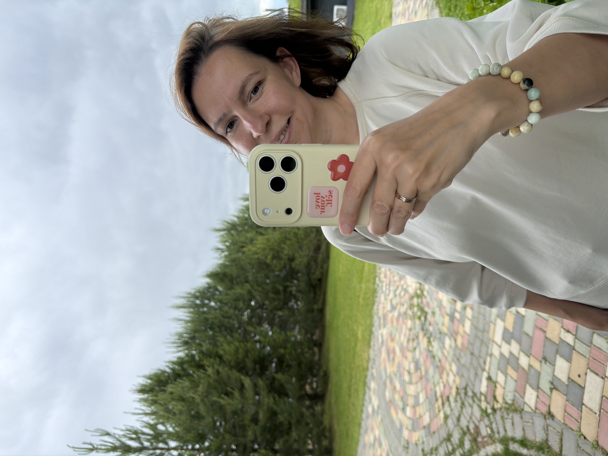 A mid-adult woman is depicted outdoors in Izhevsk, Russia, holding a light-colored smartphone in her left hand as if taking a selfie. She wears a plain gold-toned ring on her left ring finger and a bracelet composed of large, multicolored spherical beads on her right wrist. Her hair is light brown, and she displays a faint smile. The phone is encased in a light yellow/cream cover featuring a triple camera module. Two stickers adorn the phone case: a rectangular pink one with the inverted white text "love your self," and a smaller red flower sticker below it. She is wearing a white long-sleeved top. The background features a patterned paved pathway to her left, composed of multi-colored rectangular blocks in shades of red, pink, grey, cream, yellow, and brown. Beyond the pathway and behind the woman, a manicured green lawn extends to a dense line of dark green pine trees. The sky above is uniformly overcast and grey. A dark-colored building with a partially visible white vent is present in the distant right background.