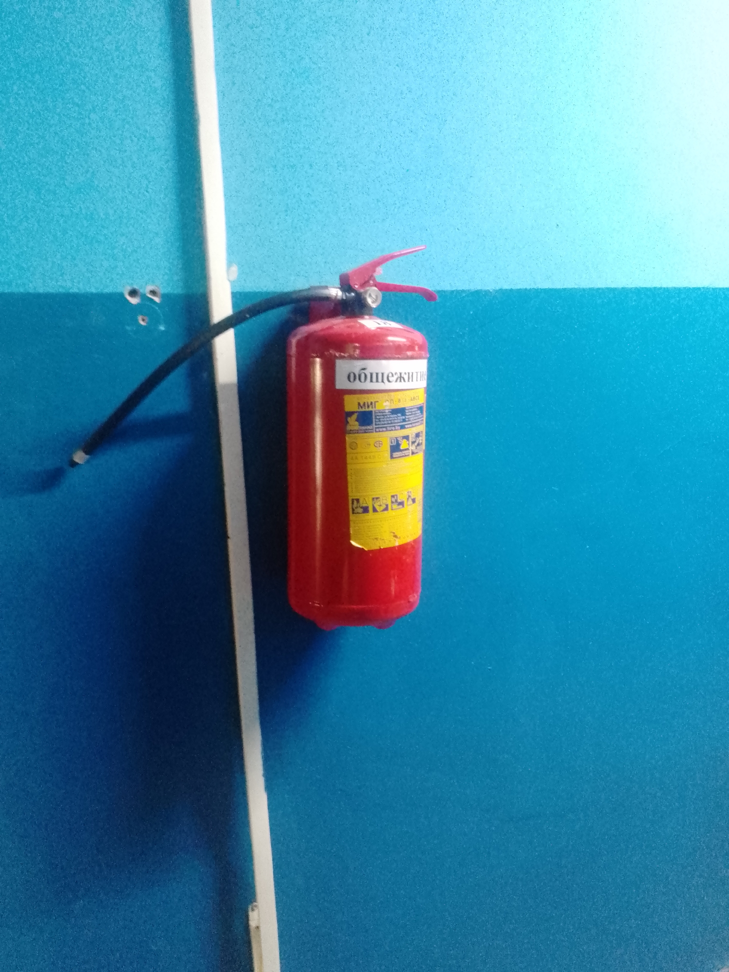 A red fire extinguisher is mounted on a blue wall. The fire extinguisher has a label with Russian text, which appears to be instructions for its use. A white vertical strip is present on the wall, to the left of the fire extinguisher. The wall has a textured surface. There are two small holes in the wall to the left of the white strip, above the fire extinguisher's hose. The overall setting is indoors, likely a public building or corridor.