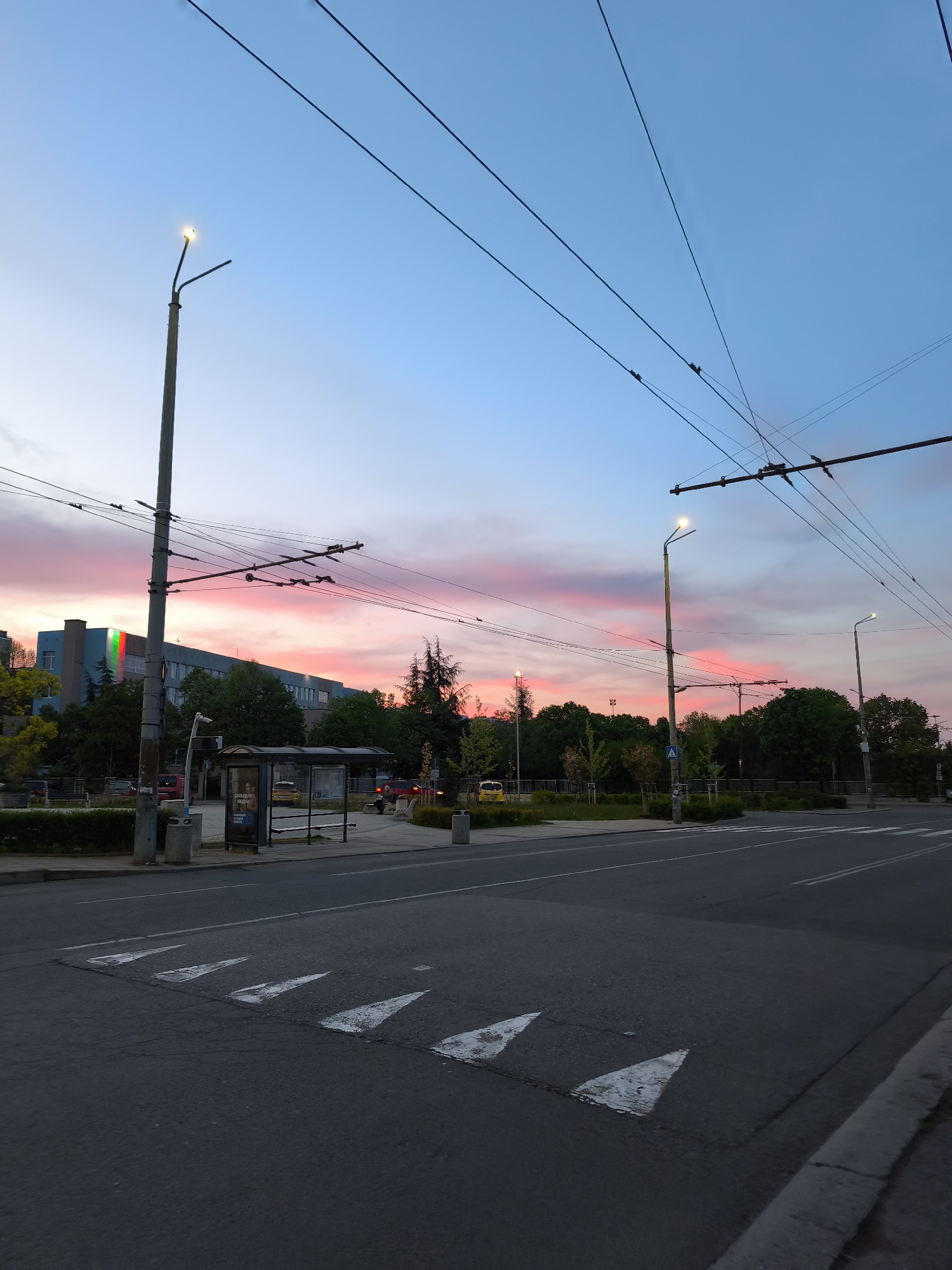 The image is a dusk scene taken on a city street in Burgas, Bulgaria. The sky is awash in colors of pink, red, and blue, with the sun having recently set or about to rise. Overhead electrical lines for trolleybuses are prominent.
On the left side of the street, there is a bus stop with a shelter, near a building with a prominent display of the Bulgarian flag, which is green, white, and red. Further down the street, a patch of green trees and park space can be seen.
On the right side of the street, there is a crosswalk with prominent triangular markings. The road is made of asphalt and shows signs of wear. Streetlights are spaced along the road, emitting a warm, yellow light. There appears to be a person at the bus stop to the left.