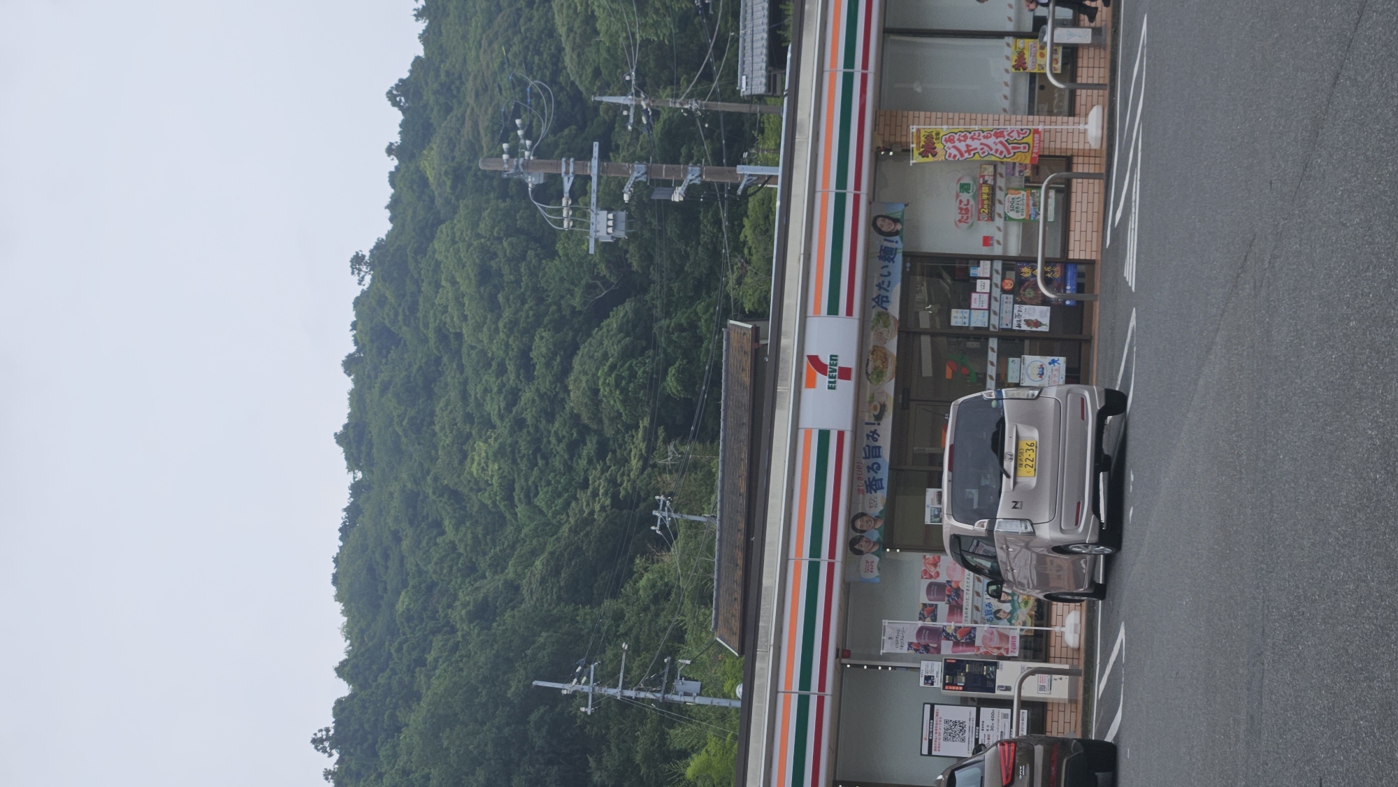 A tan 7-Eleven convenience store is situated at the base of a densely forested hill in Kamakura, Japan. The store has a prominent red and white "7-ELEVEN" logo. In front of the store, a light-colored, compact car with a yellow license plate is parked. To the left of this car, another vehicle, partially visible, is also parked. The parking lot surface is asphalt. The sky is overcast. Utility poles and electrical wires are visible in front of the wooded hillside.