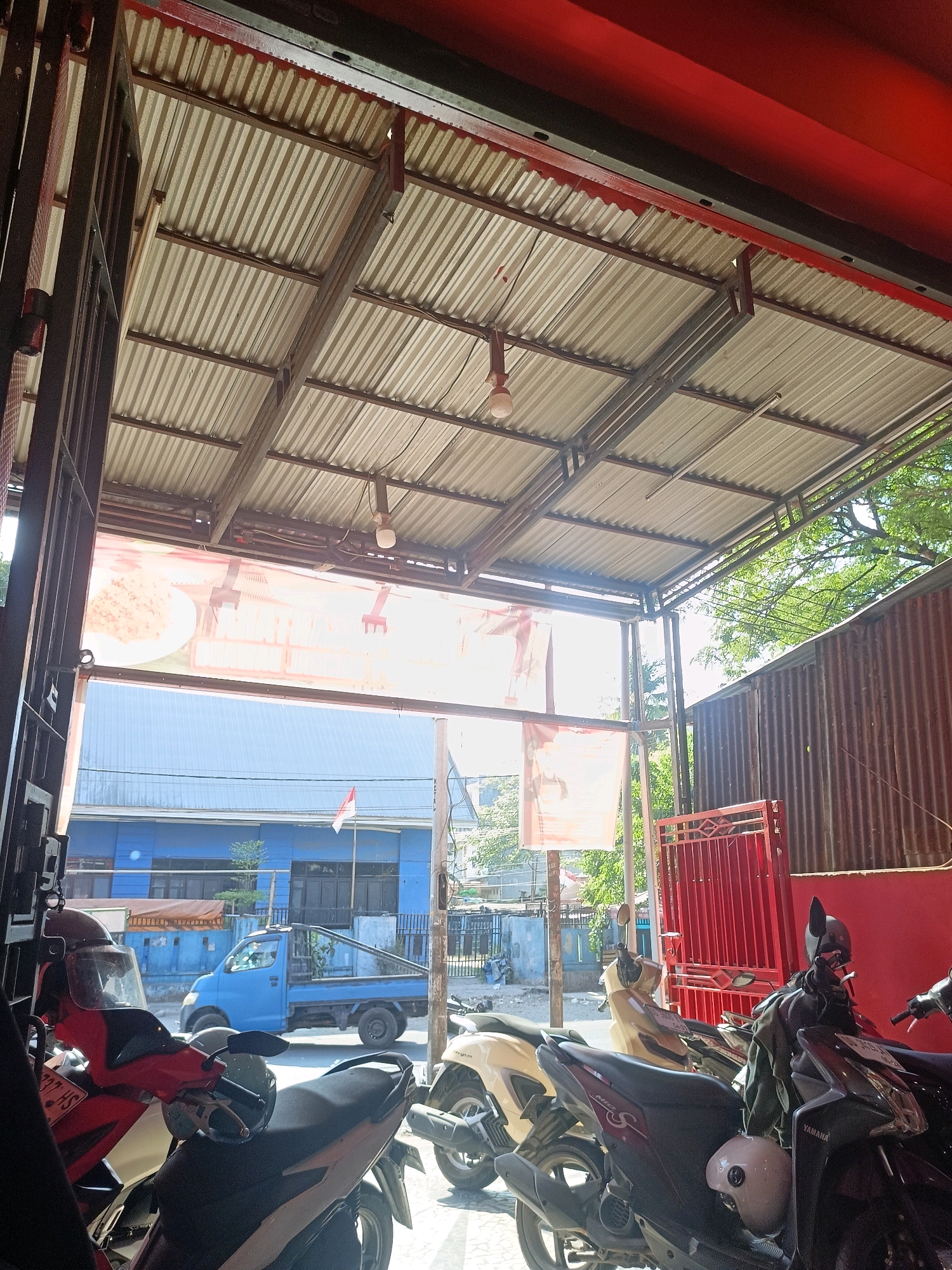 The image captures a street-level view of a covered outdoor area, likely a parking or waiting area, adjacent to a building. In the foreground, several parked motorcycles are visible, including a red and white scooter on the left and a dark-colored scooter on the right with "YAMAHA" branding. Behind the motorcycles, a blue truck is parked, and a red metal gate is seen. The background reveals a blue building with windows and a red and white flag flying. Above, a corrugated metal roof supported by metal beams covers the area. Trees and foliage are visible in the distance, suggesting a semi-urban or suburban environment. The scene appears to be in Makassar, Indonesia, given the presence of the Indonesian flag.