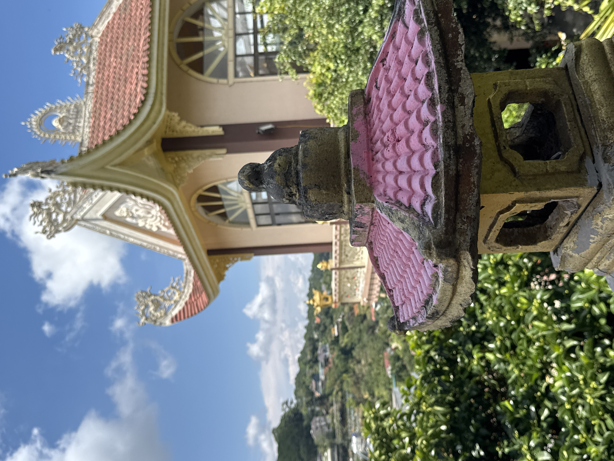 The image depicts a close-up view of a weathered, pink-tiled pagoda lantern in the foreground, set against a backdrop of lush greenery and a light-colored building with traditional Asian architecture, possibly a temple or pagoda. The building features a red-tiled roof with ornate golden trim and decorative elements. In the distance, rolling hills are visible, dotted with houses and trees, suggesting a hillside location. The sky is blue with scattered white clouds. This scene is characteristic of the picturesque landscape and spiritual sites found in Da Lat, Vietnam, a city known for its French colonial architecture, verdant hills, and numerous pagodas.