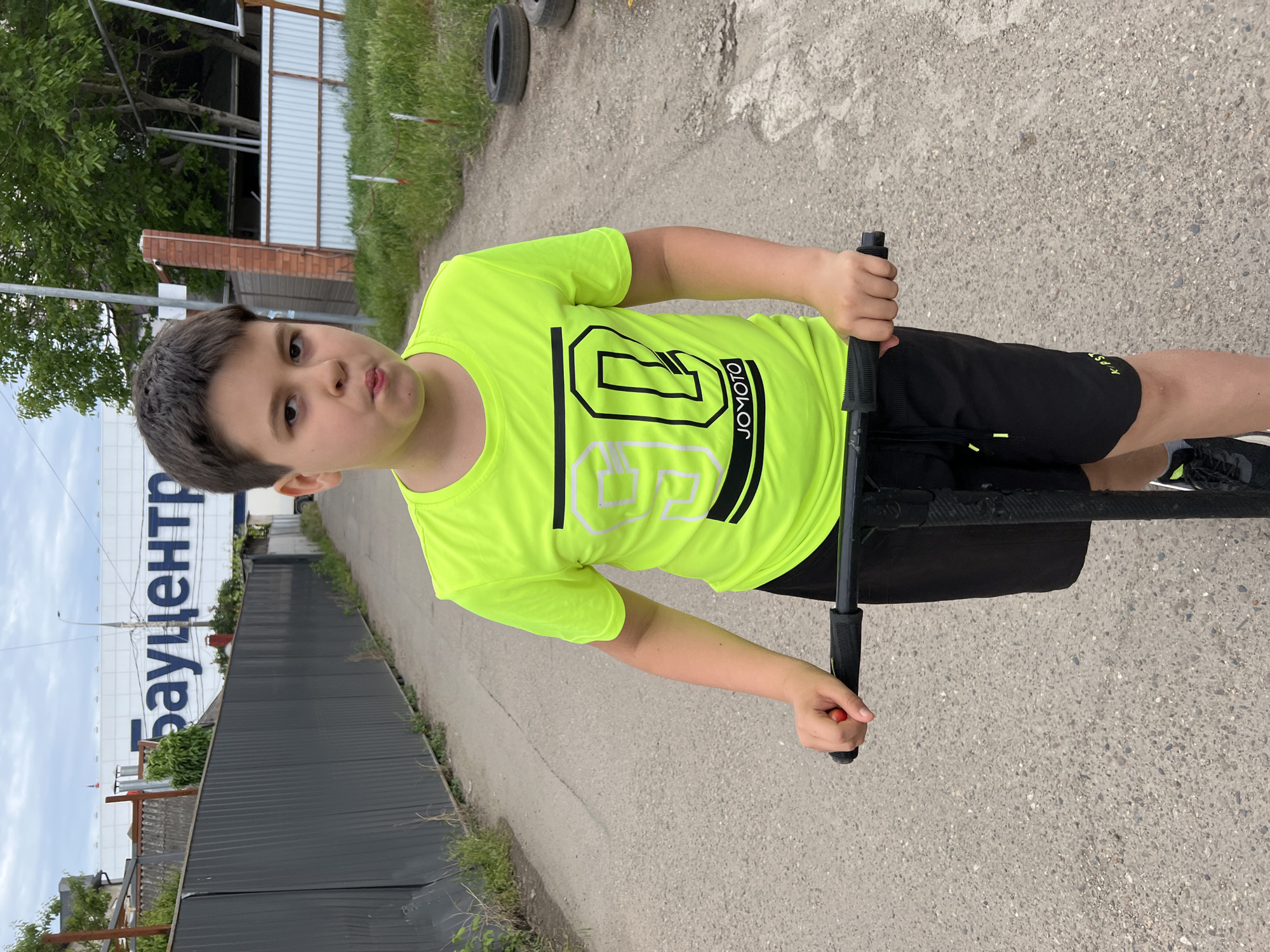 A child in a bright yellow shirt is standing on a pogo stick on an asphalt surface. The background includes a gray metal fence, foliage, and a building with the word "Бауцентр" visible, indicating the location is Krasnodar, Russia.