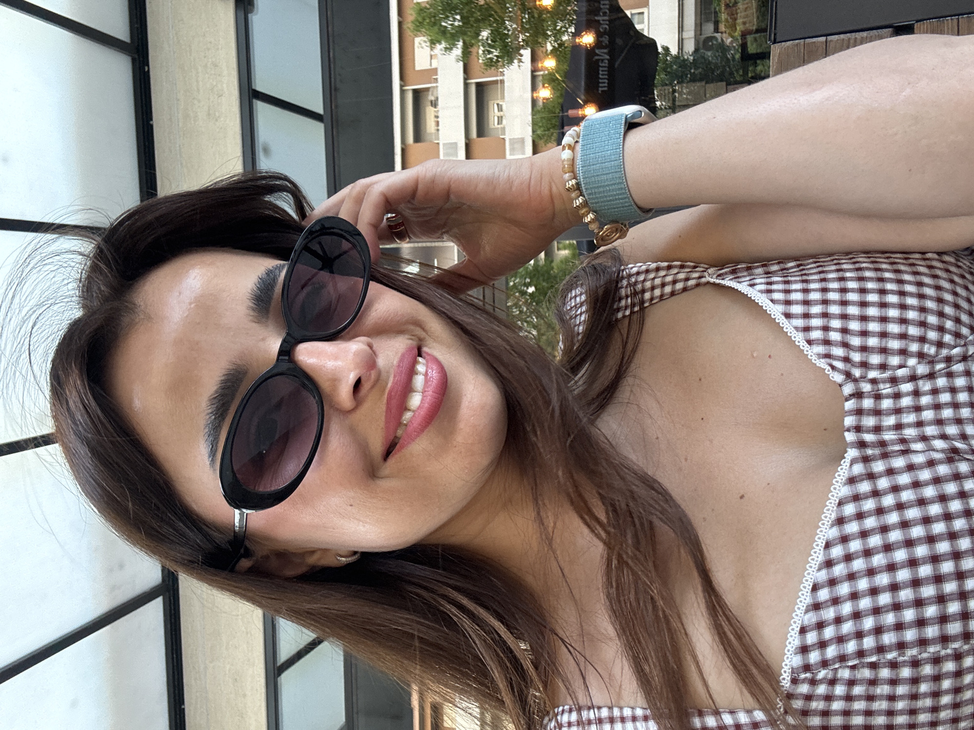 A woman with long, dark brown hair and fair skin is shown in a close-up, selfie-style shot outdoors. She is wearing oval-shaped black sunglasses and has a bright pink lipstick on her lips. Her right hand is raised to adjust her sunglasses, revealing a ring and a bracelet with blue and beige beads on her wrist. She is wearing a white and maroon gingham-print top with white lace trim. In the background, there are glimpses of a building with balconies, trees, and string lights, suggesting an urban outdoor setting. The location is specified as Podgorica, Montenegro.